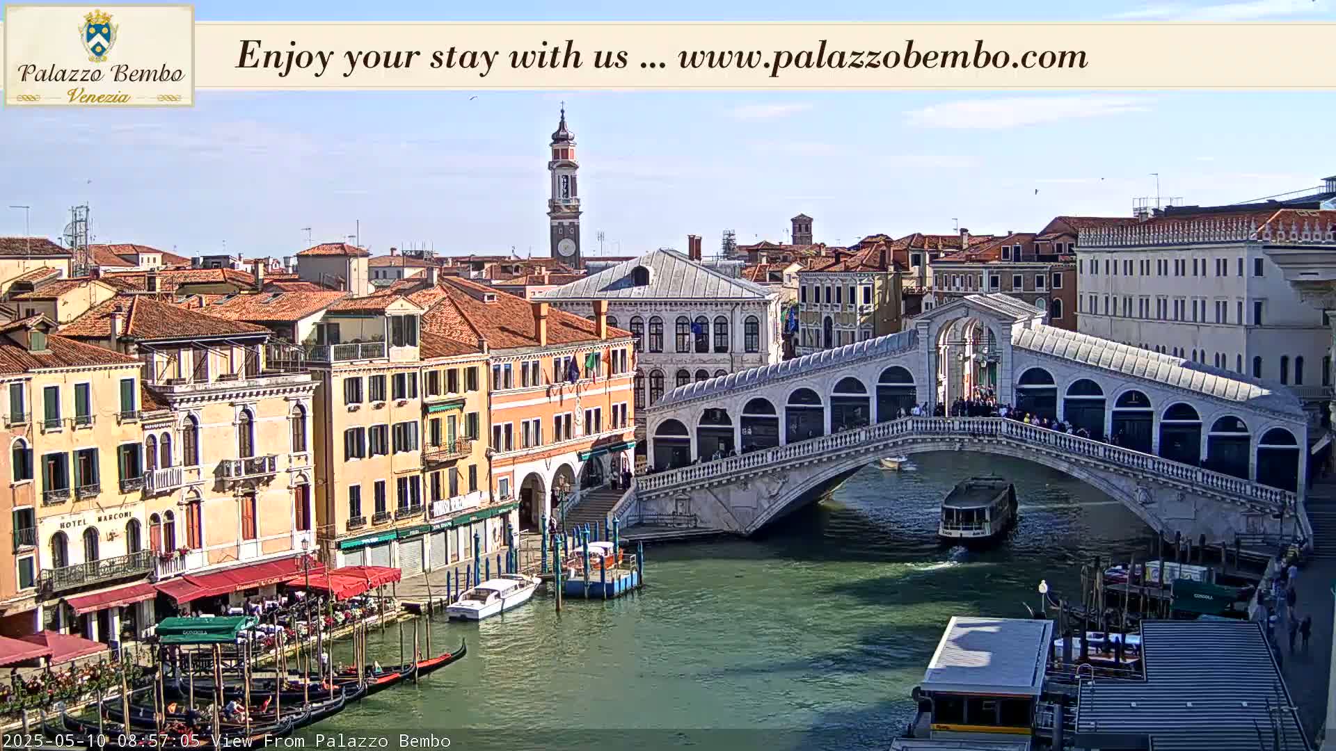 A sunny view of a canal in Venice, with gondolas and a bridge filled with people.