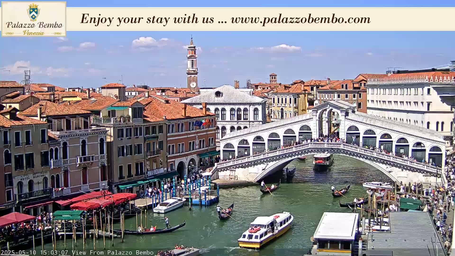 A sunny day in Venice shows a canal with gondolas and boats passing under the Rialto Bridge, which is crowded with people.