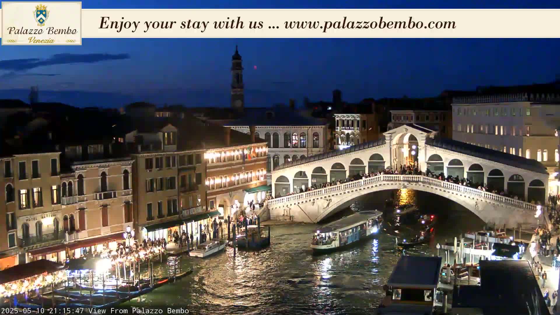 A nighttime view of a Venetian canal with a bridge crowded with people, boats on the water, and illuminated buildings lining the waterway under a dark blue sky.