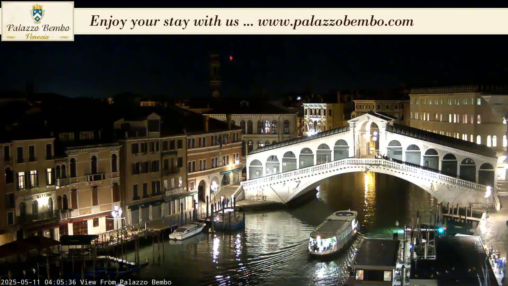 A nighttime view of a lit bridge spanning a canal in Venice, with boats on the water and buildings illuminated along the waterway.