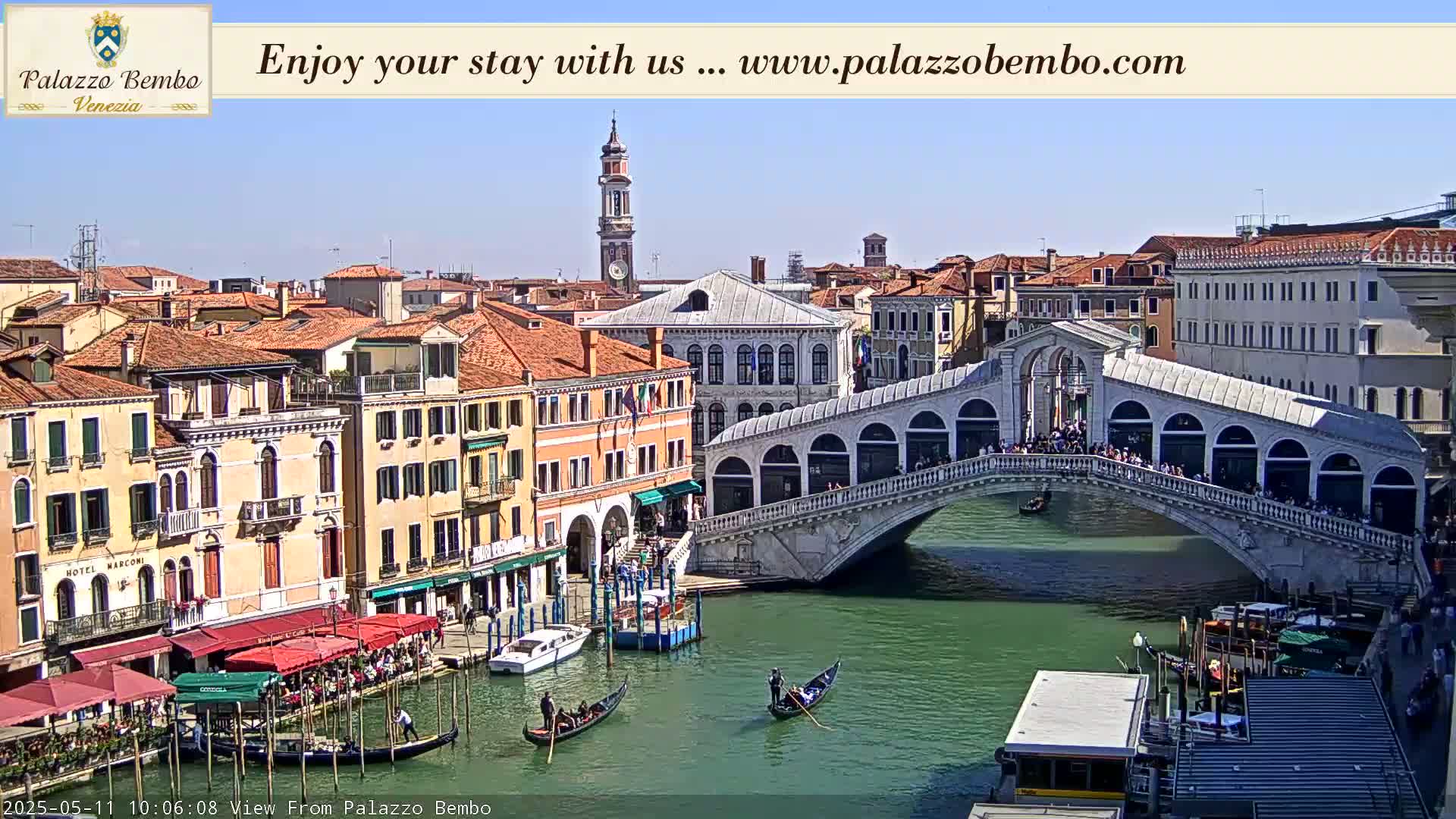A sunny view of a canal in Venice, showing a bridge crowded with people, gondolas on the water, and buildings lining the waterway.