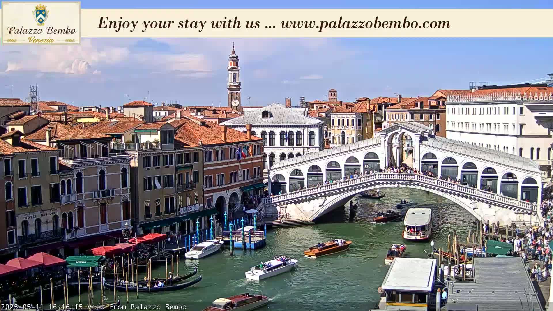 A sunny day in Venice shows a canal with boats, the Rialto Bridge crowded with people, and many buildings lining the waterway.