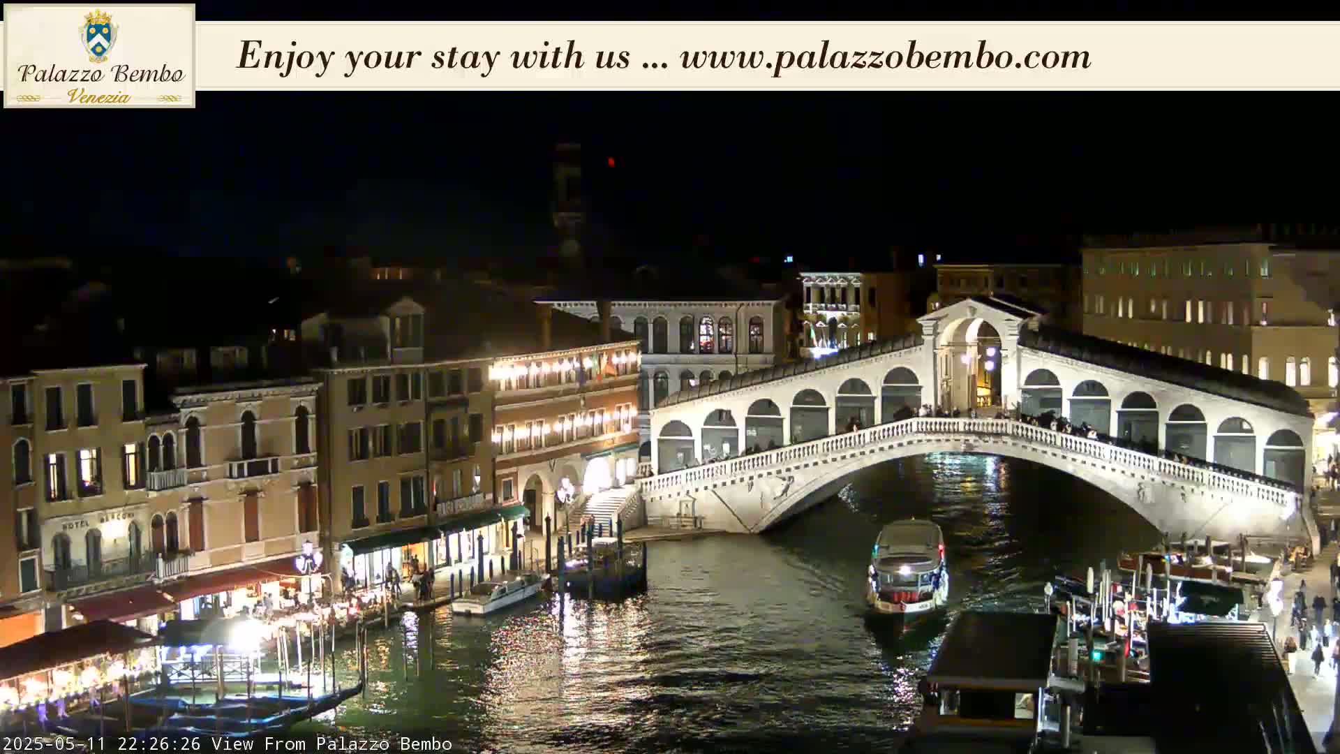 A nighttime view of a canal in Venice shows a bridge lit up with people on it and boats passing underneath.