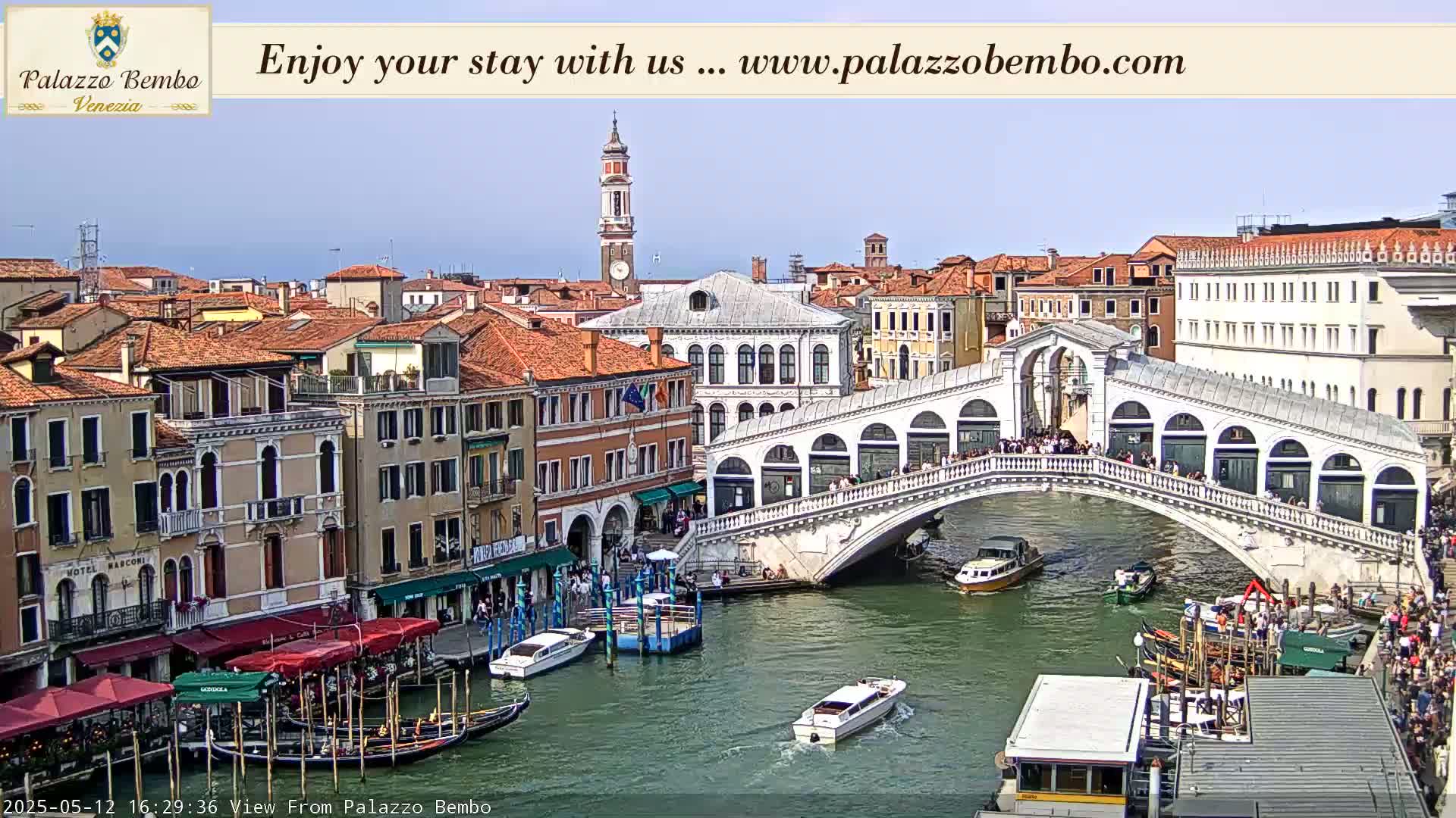 A sunny view of a canal in Venice, Italy, shows a bridge crowded with people, boats on the water, and buildings lining the waterway.