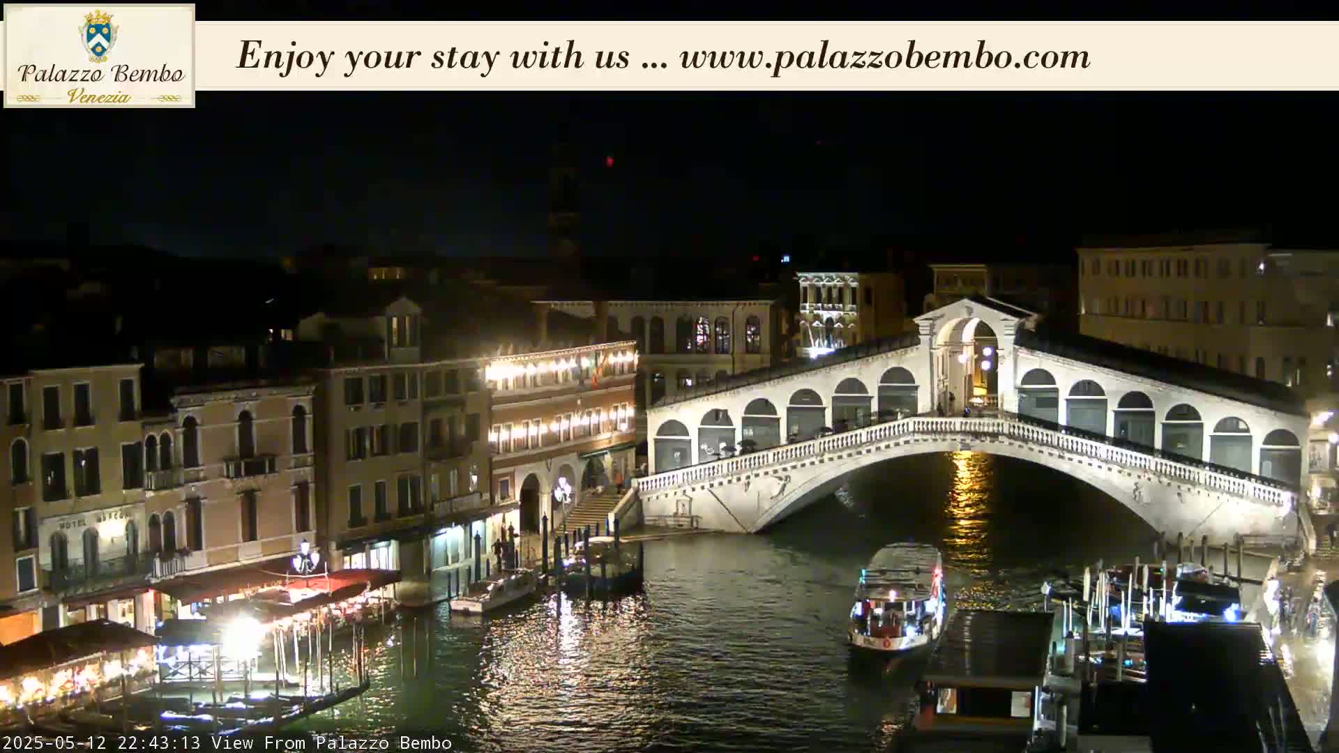A nighttime view of a lit-up bridge over a canal in Venice, with buildings and boats along the waterway.