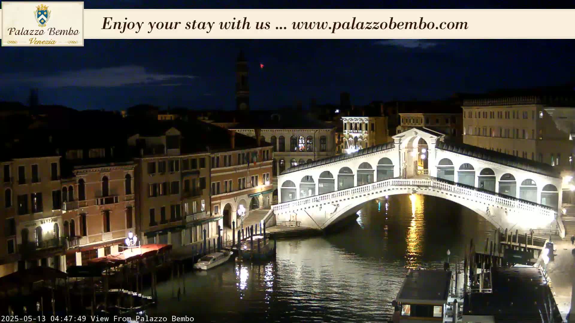 A nighttime view of a canal in Venice, showing a lit bridge and buildings along the waterway under a dark sky.