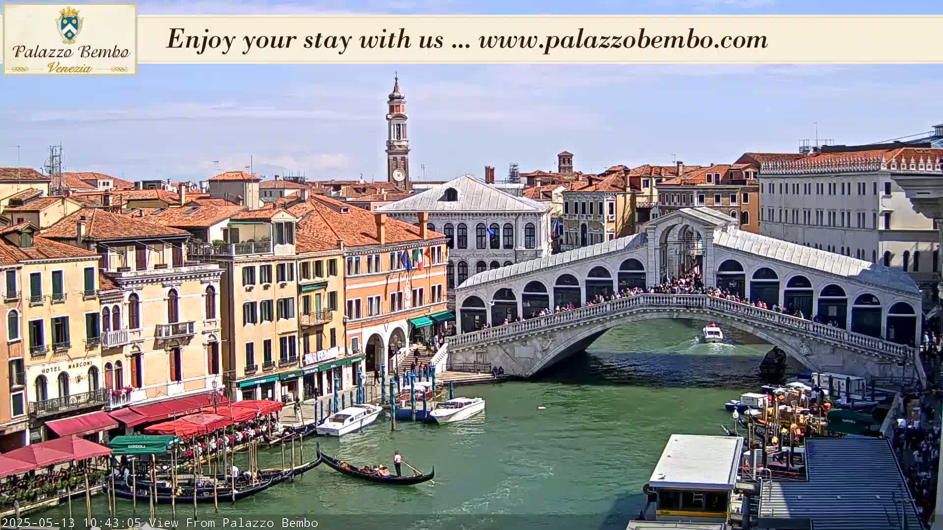 A sunny view of a canal in Venice, showing a bridge crowded with people, gondolas, and boats, with buildings lining the waterway.
