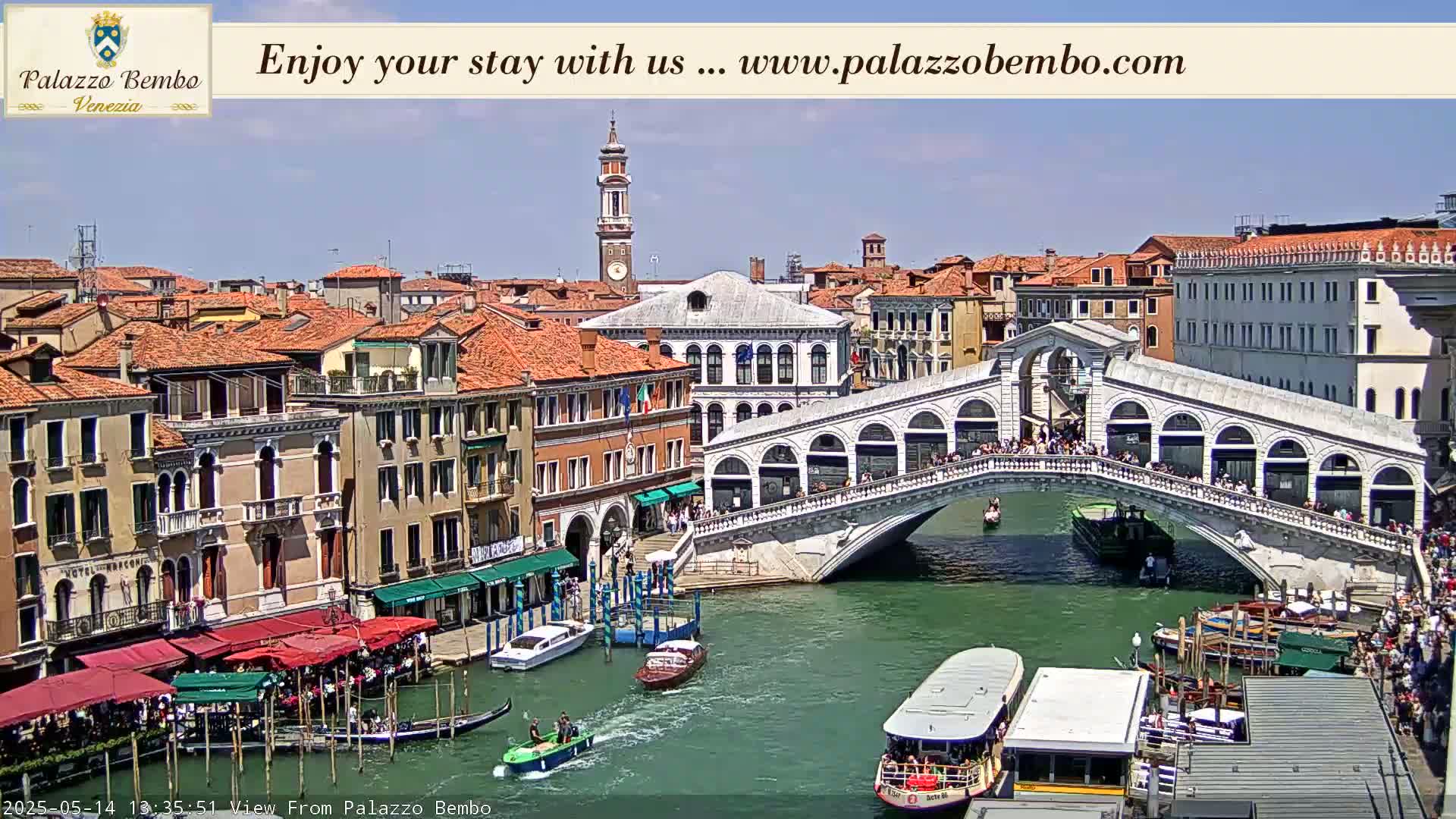 A sunny day in Venice shows the Rialto Bridge crowded with people, boats on the canal below, and colorful buildings lining the waterway.