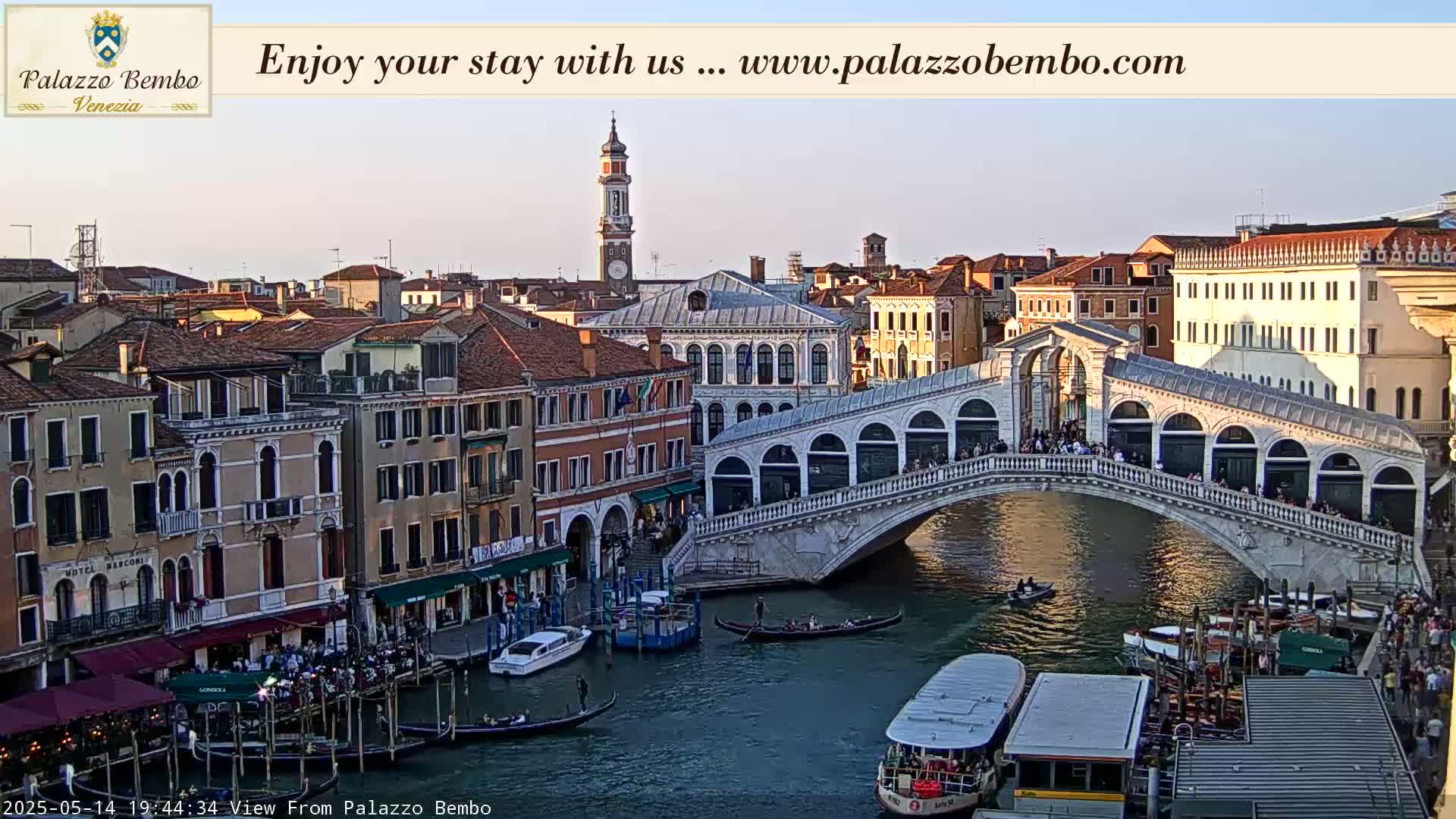 A Venetian bridge arches over a canal, with gondolas and boats below, under a twilight sky.
