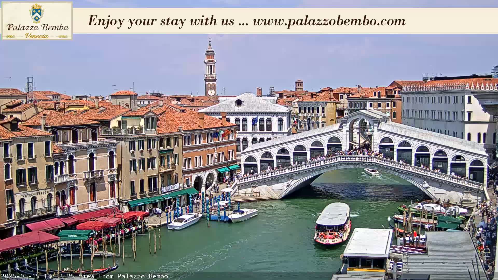A sunny view of a canal in Venice, Italy, shows a bridge crowded with people, boats moored along the canal's edge, and buildings lining the waterway.