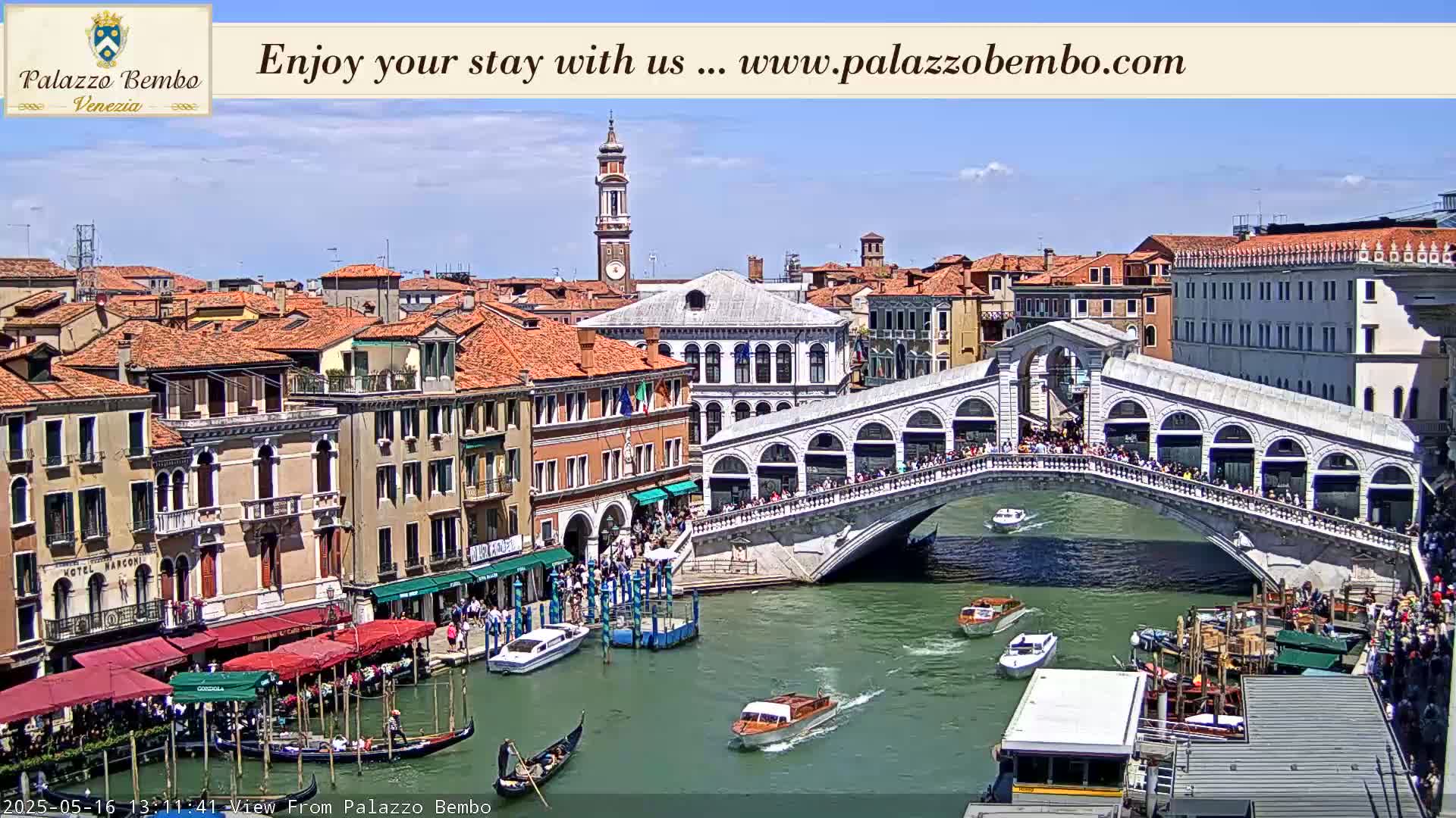 A sunny day in Venice shows a canal with boats and gondolas passing under a bridge crowded with people, with buildings lining the waterway.