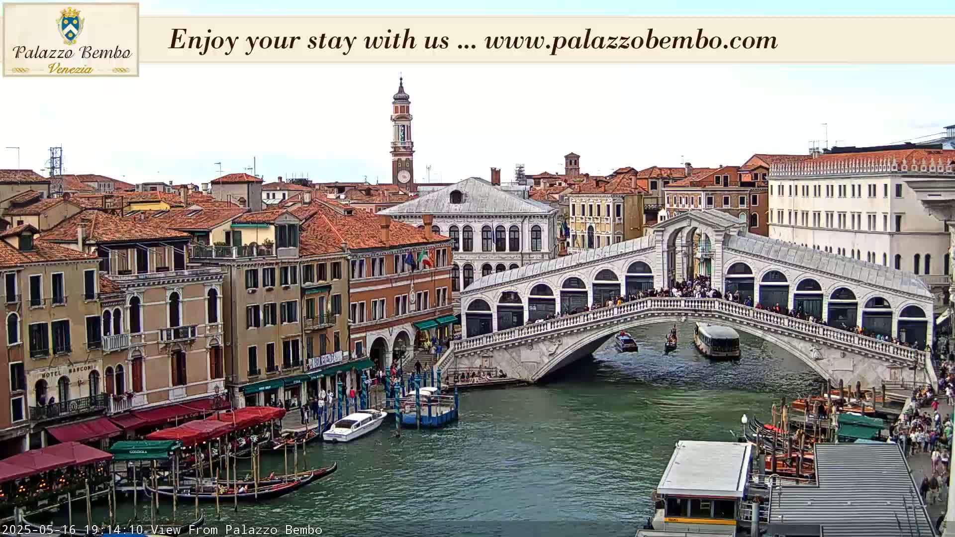 A view of a Venetian canal shows a bridge with people walking across it, gondolas and other boats on the water, and several buildings lining the canal under a mostly cloudy sky.