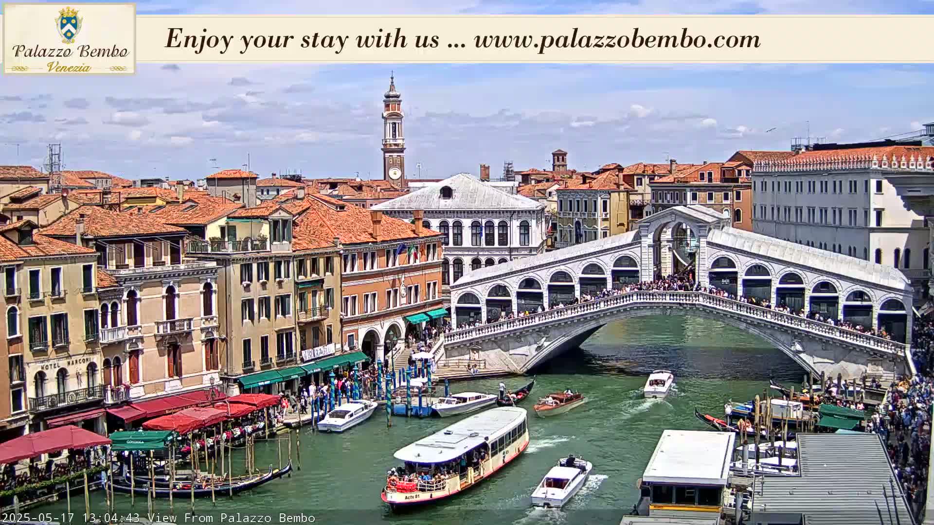 A sunny day in Venice shows a canal with boats, the Rialto Bridge crowded with people, and numerous buildings lining the waterway.