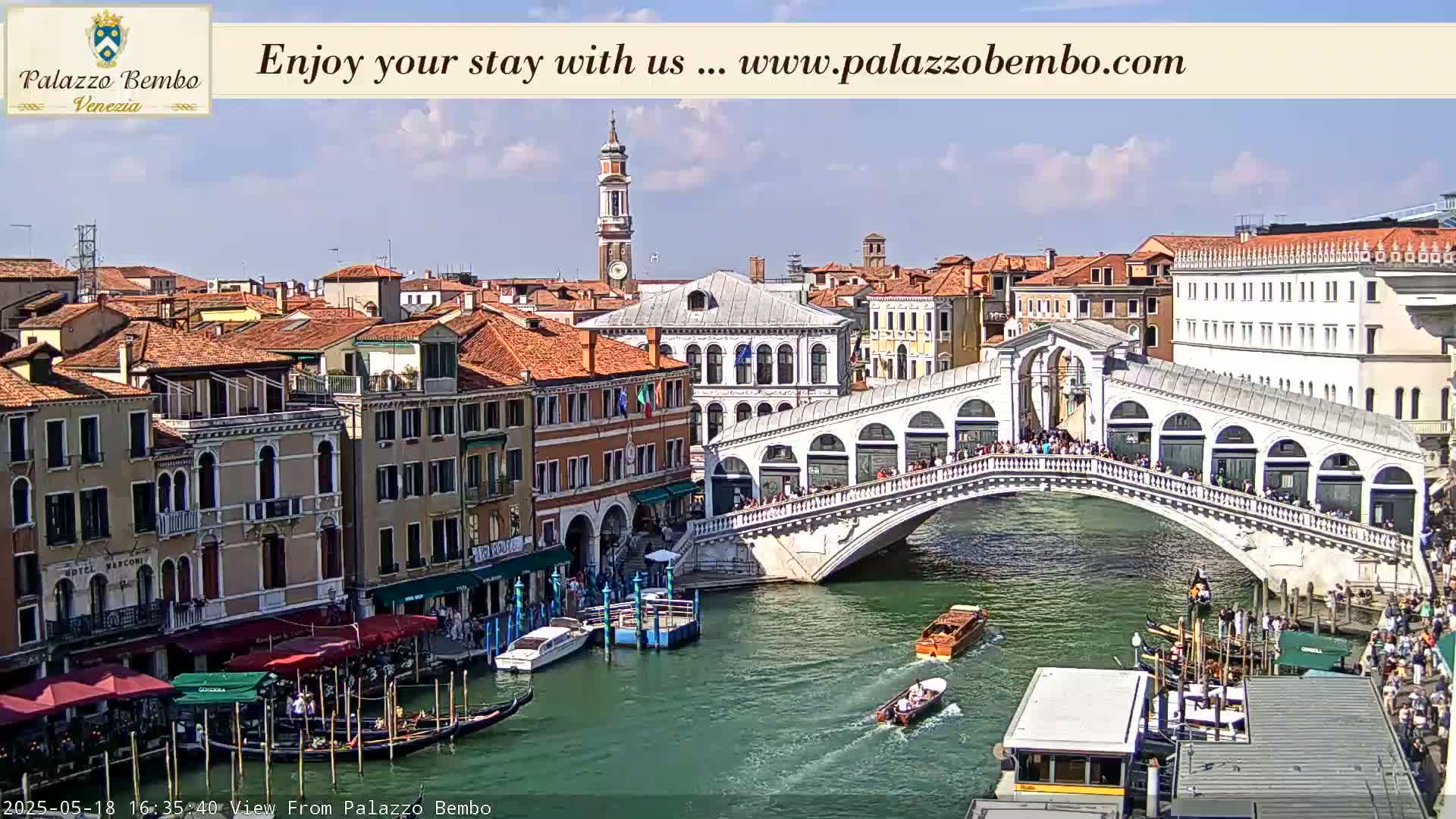 A sunny view of a canal in Venice, Italy, shows a bridge crowded with people, boats on the water, and buildings lining the waterway.
