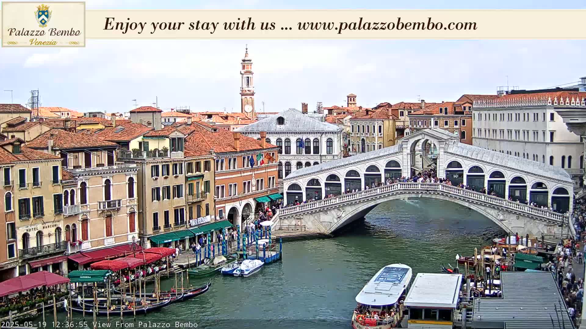 A Venetian canal scene on a partly cloudy day shows a bridge crowded with people, boats moored along the canal's edges, and buildings lining the waterway.