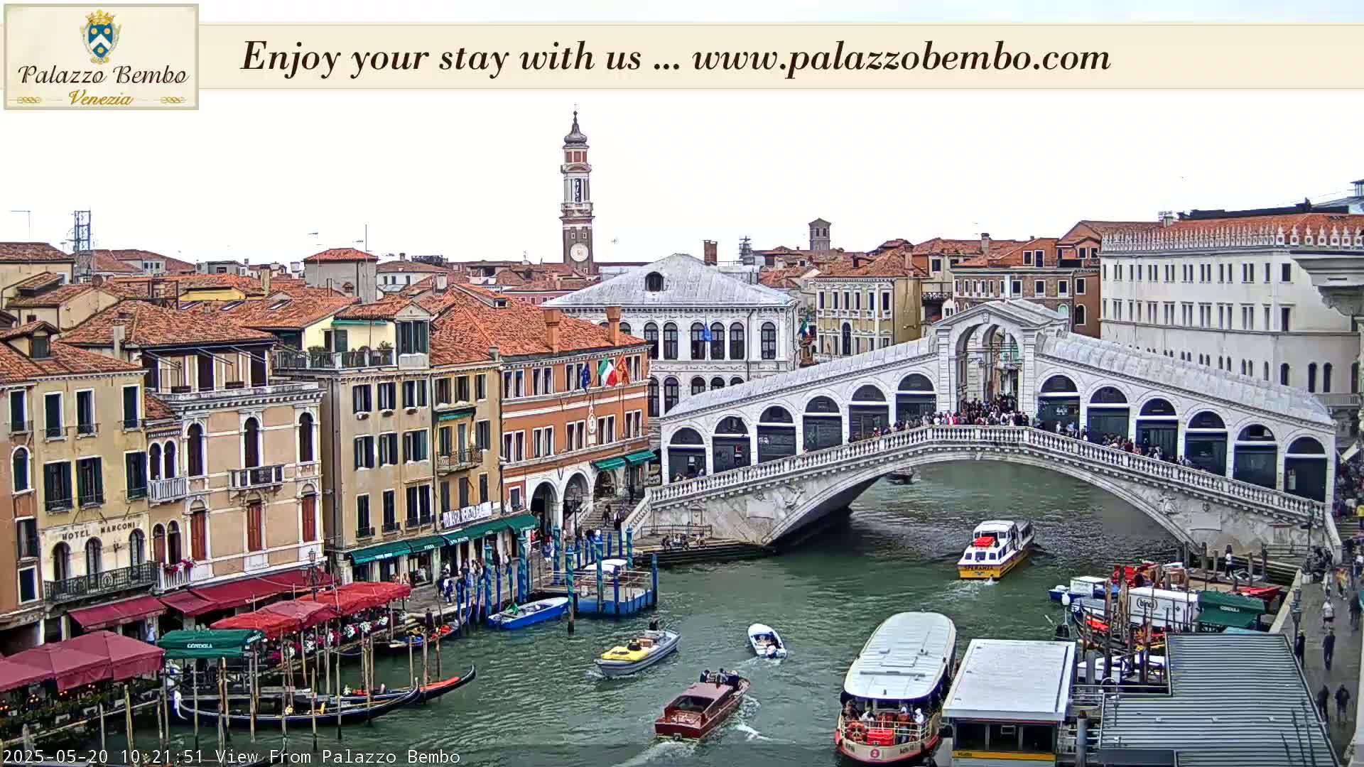A canal in Venice, under an overcast sky, features the Rialto Bridge with pedestrians crossing it and numerous boats, gondolas, and vaporetti navigating the waterway, with buildings lining the canal's edges.