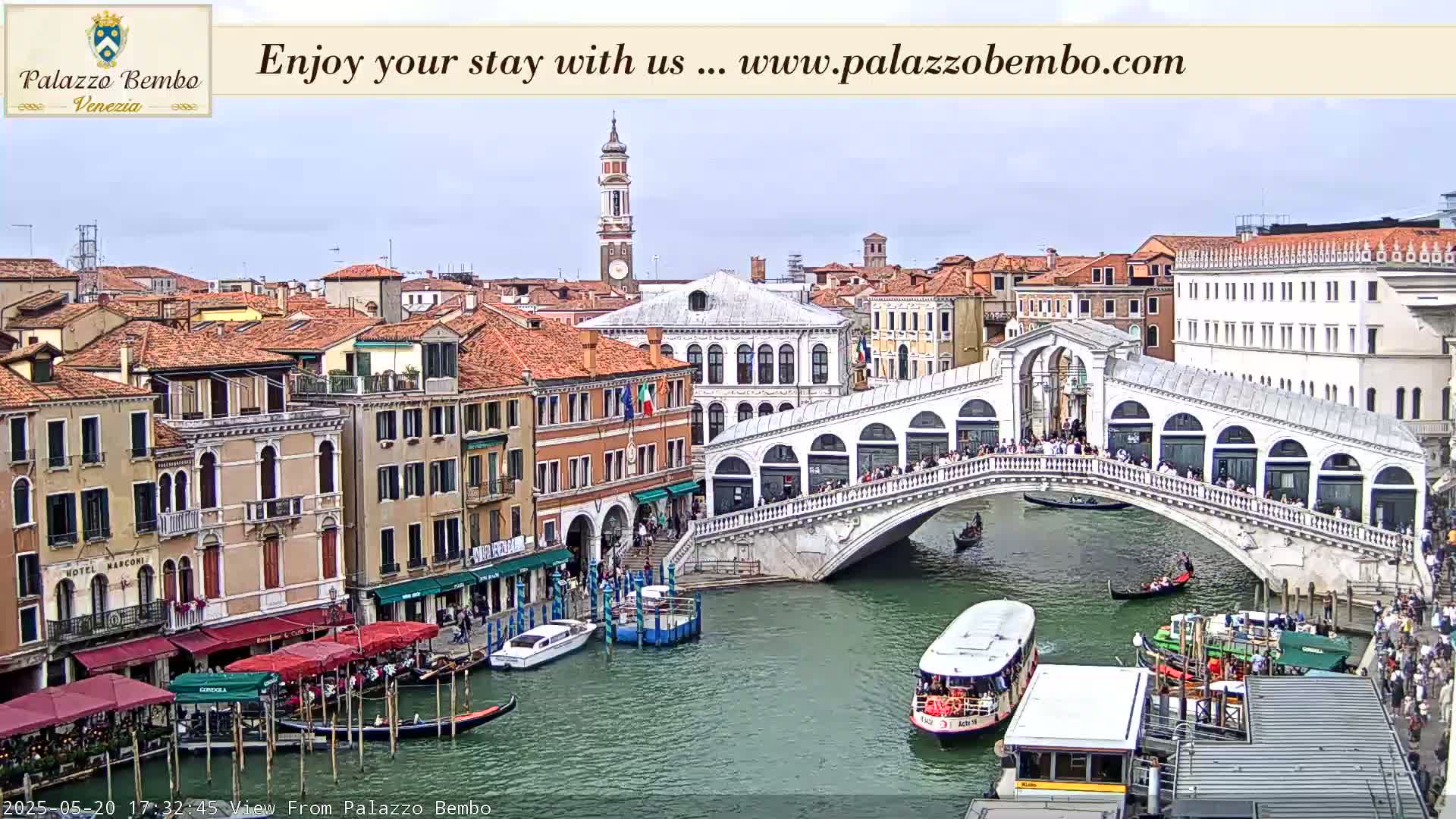 A view of a canal in Venice, under a mostly cloudy sky, shows a bridge with people walking across it and boats on the water.