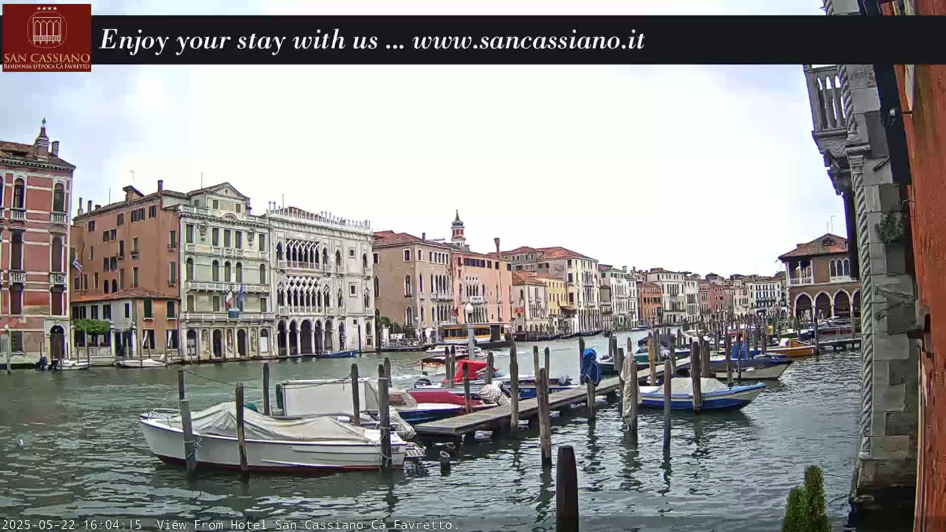 A canal in Venice, Italy, is shown on an overcast day, with numerous boats moored along a wooden dock in front of colorful buildings.