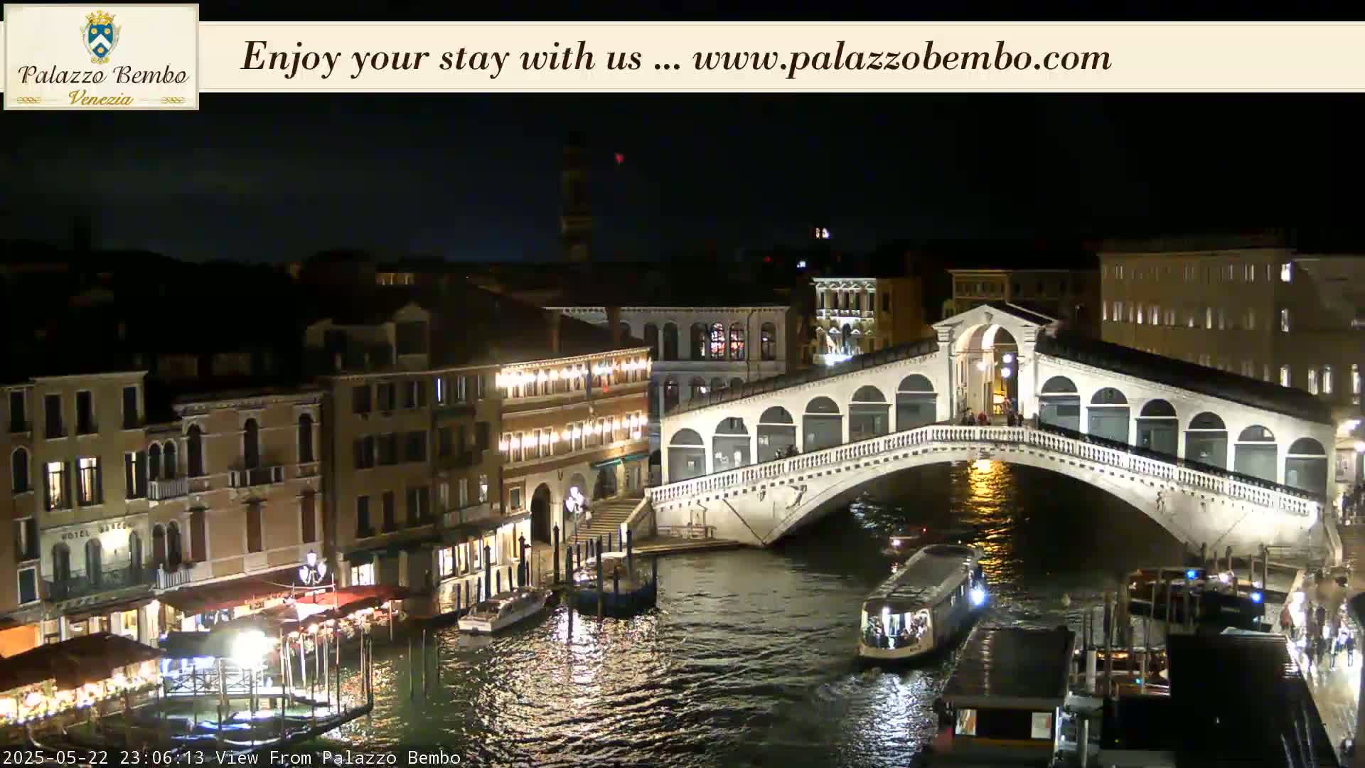 A nighttime view of a canal in Venice, showing a bridge lit up with lights, boats on the water, and buildings along the waterway.