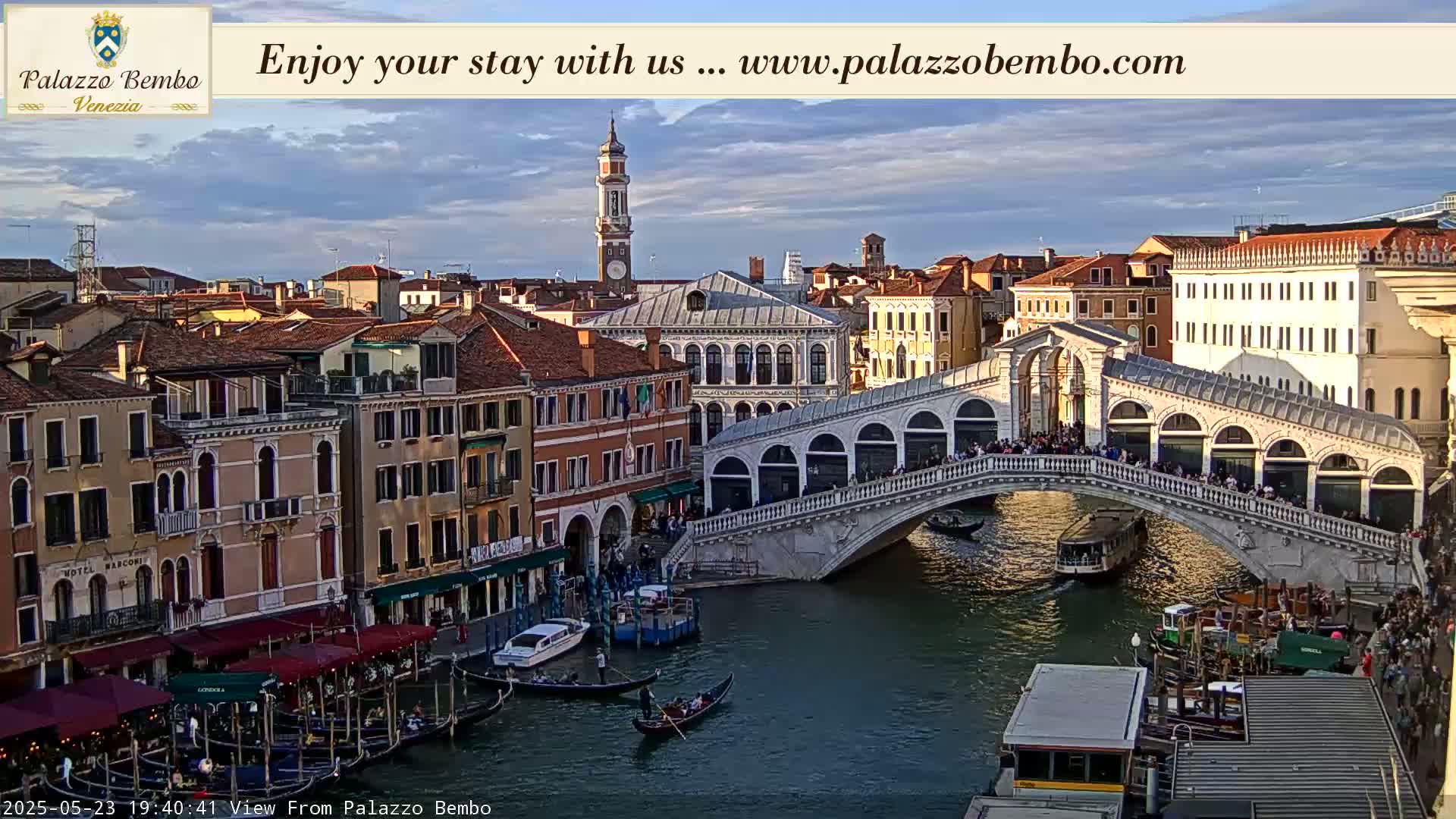 A Venetian canal scene under a partly cloudy sky shows a bridge crowded with people, gondolas, and water traffic.