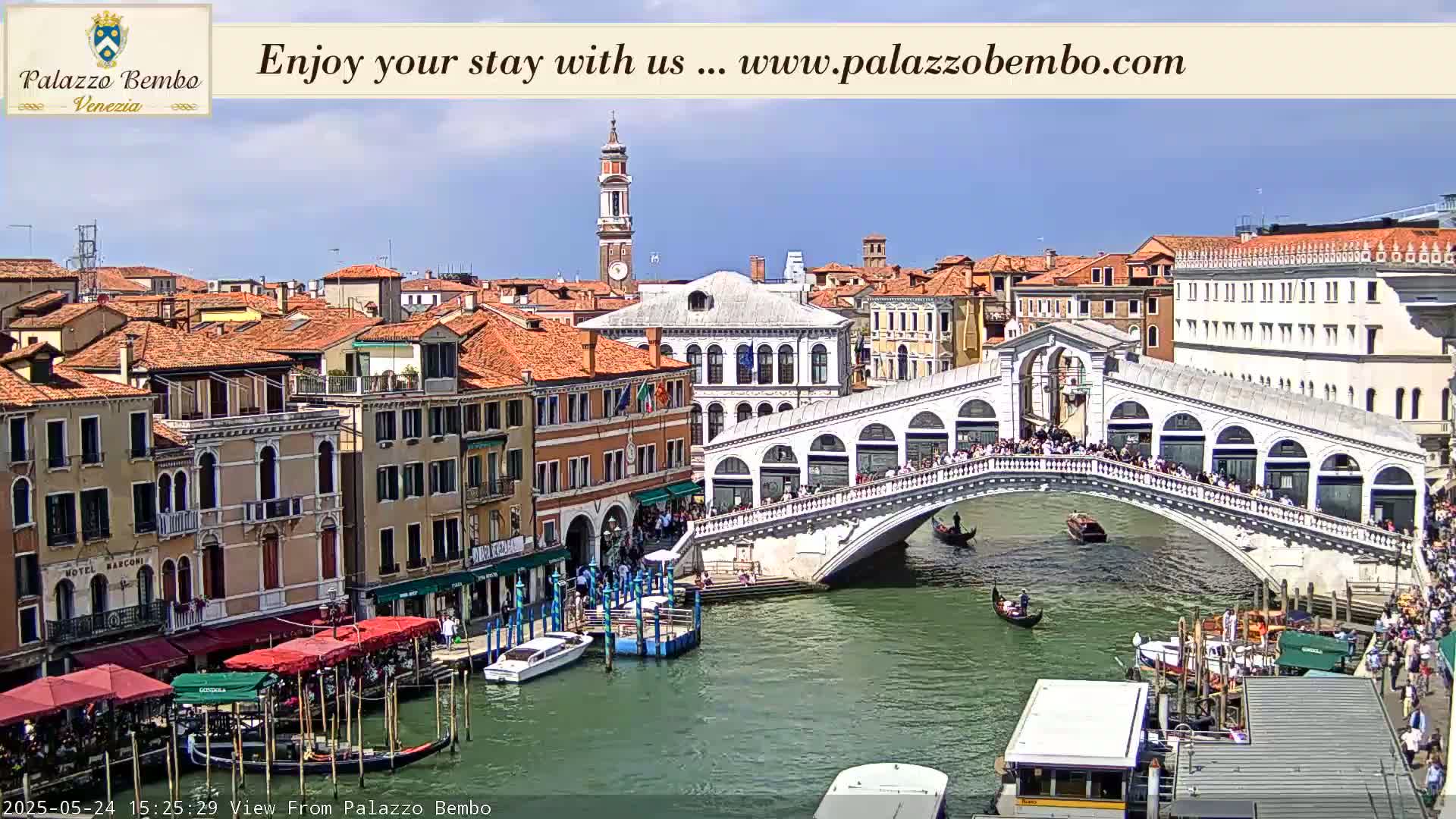 A sunny view of a canal in Venice, showing the Rialto Bridge crowded with people and gondolas on the water.