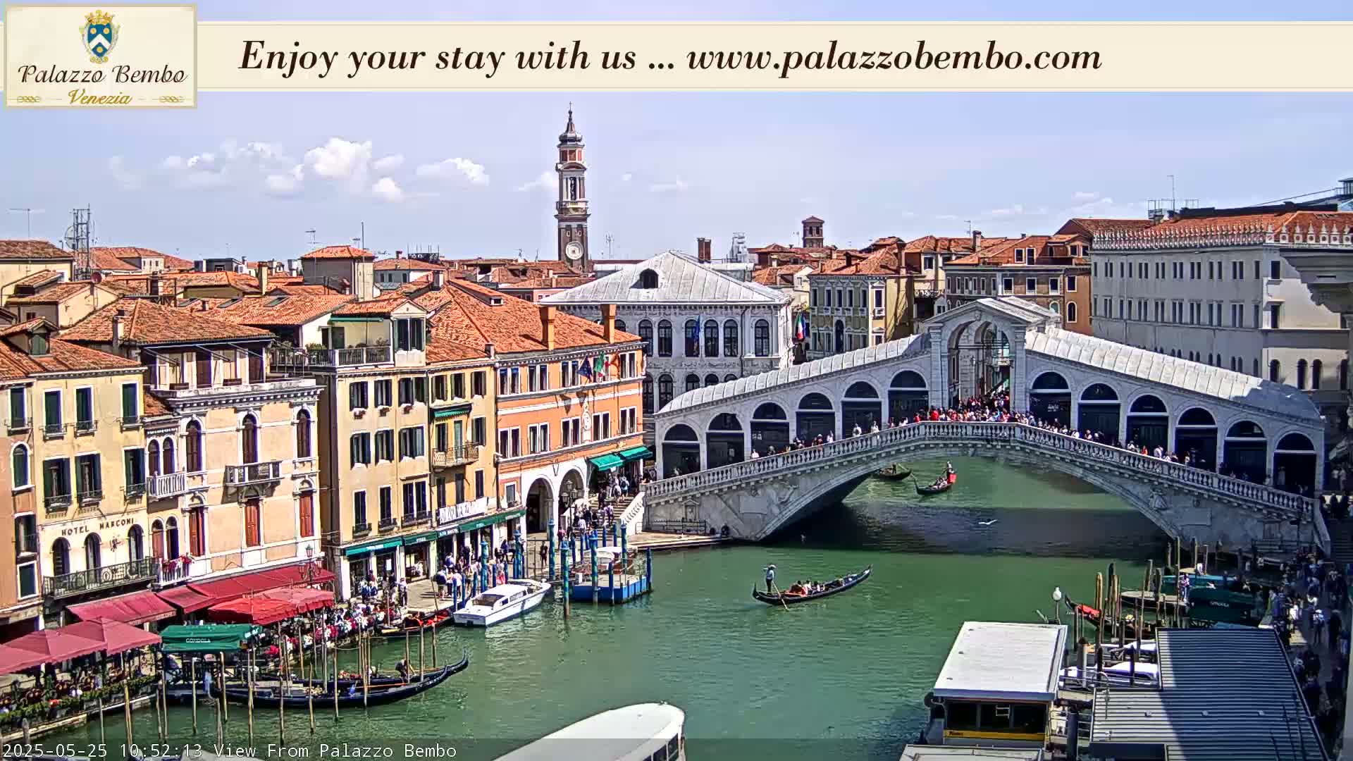 A sunny day in Venice shows a canal with gondolas and a stone bridge crowded with people, surrounded by buildings.