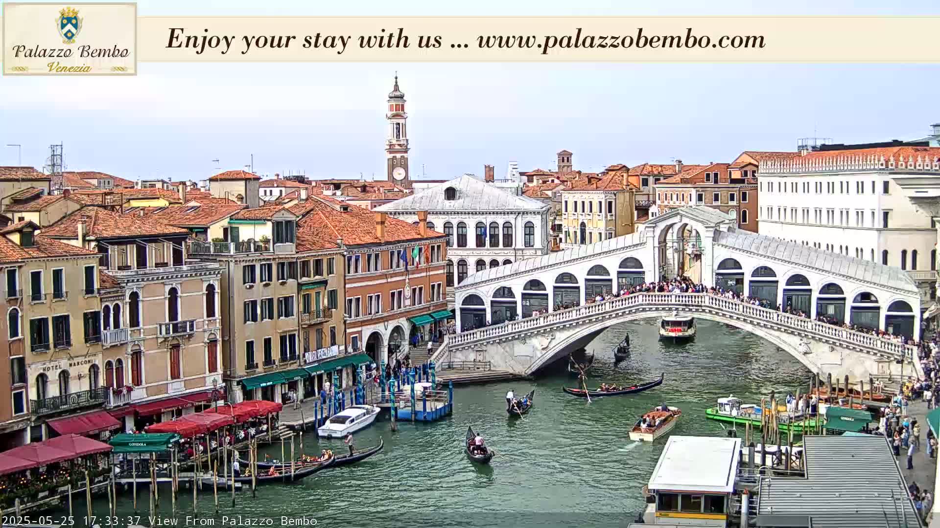 A canal in Venice, under a mostly clear sky, is filled with gondolas and boats, with the Rialto Bridge crowded with people in the background.