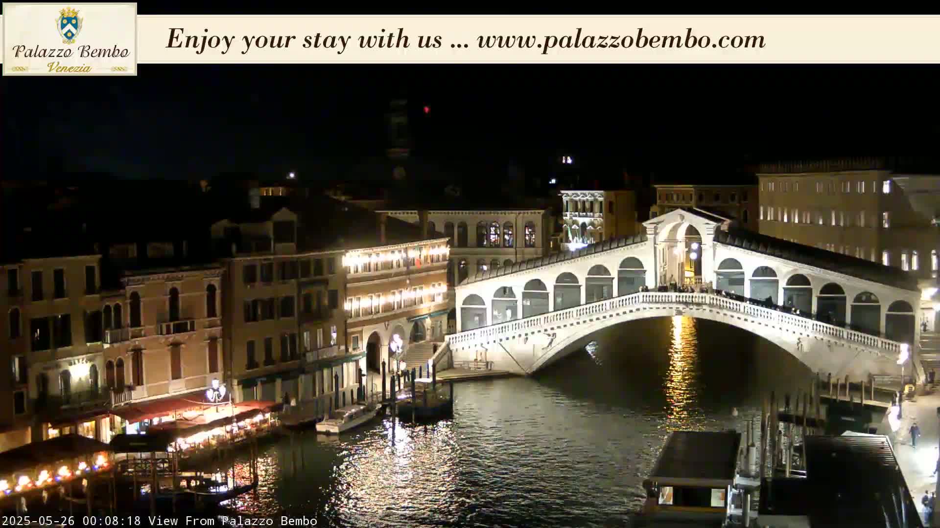 A nighttime view of a canal in Venice, showing a bridge lit up with lights and buildings along the water's edge.