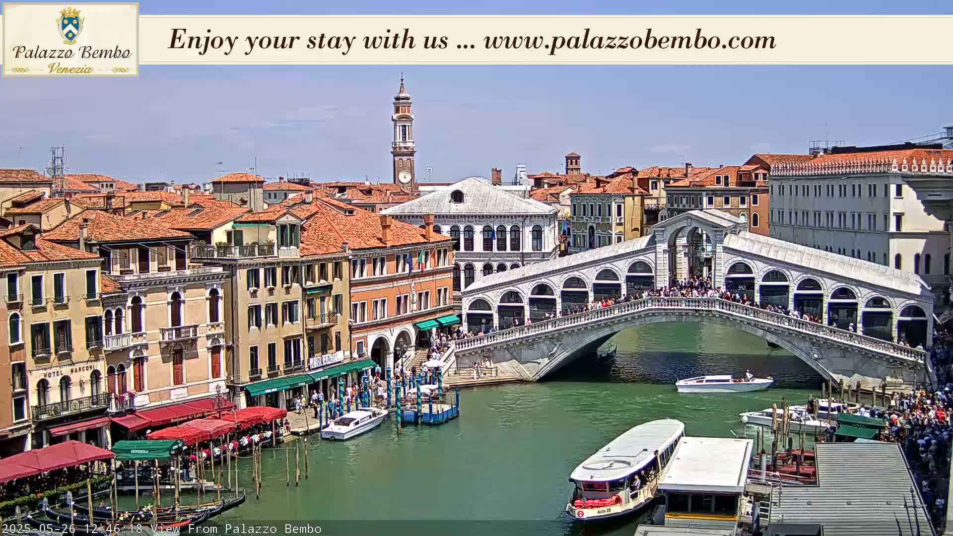 A sunny day in Venice shows a canal with boats, a crowded bridge, and many buildings with orange-tiled roofs.