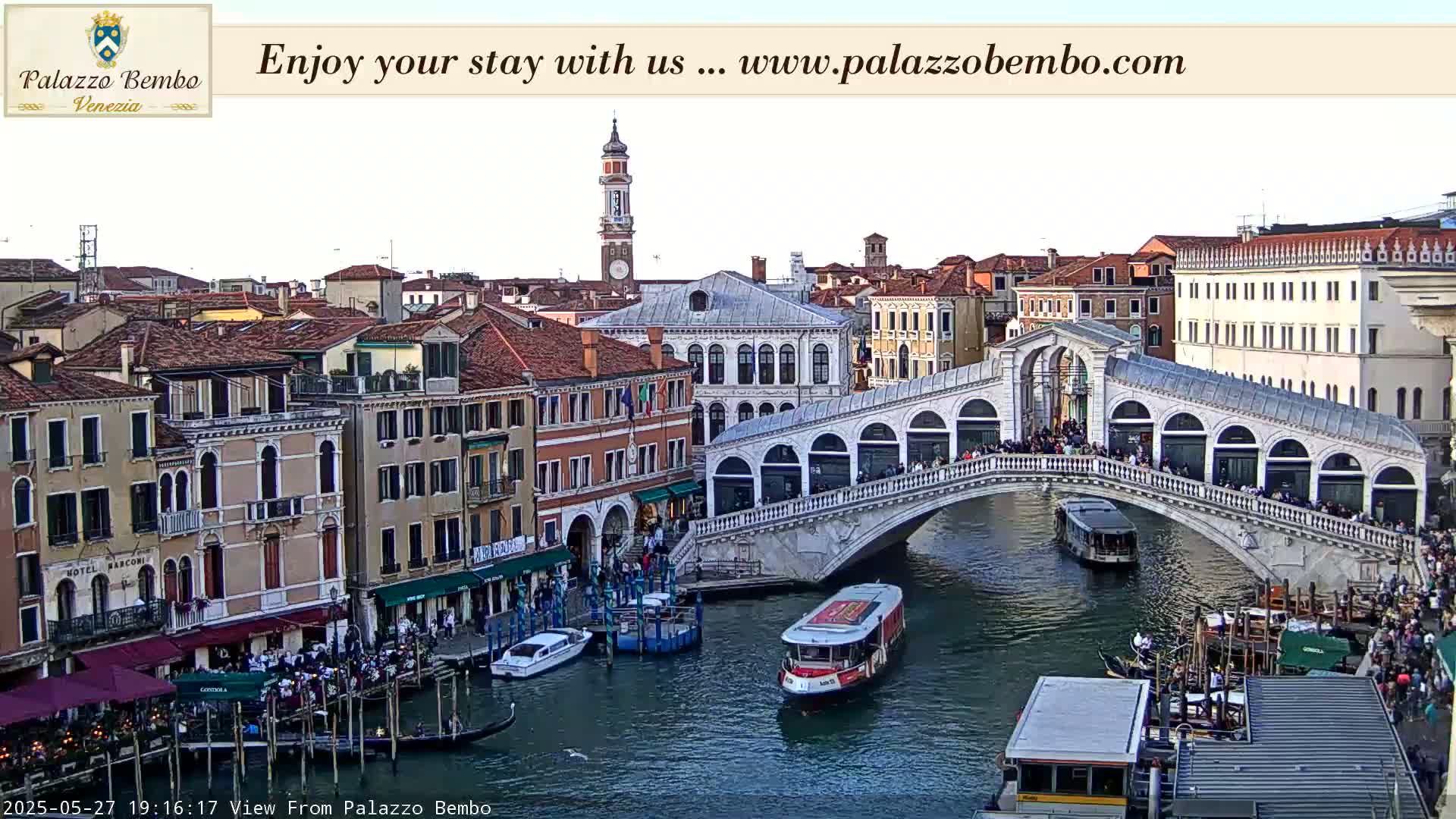 A Venetian bridge spans a canal lined with buildings, gondolas, and water taxis under a mostly clear sky.