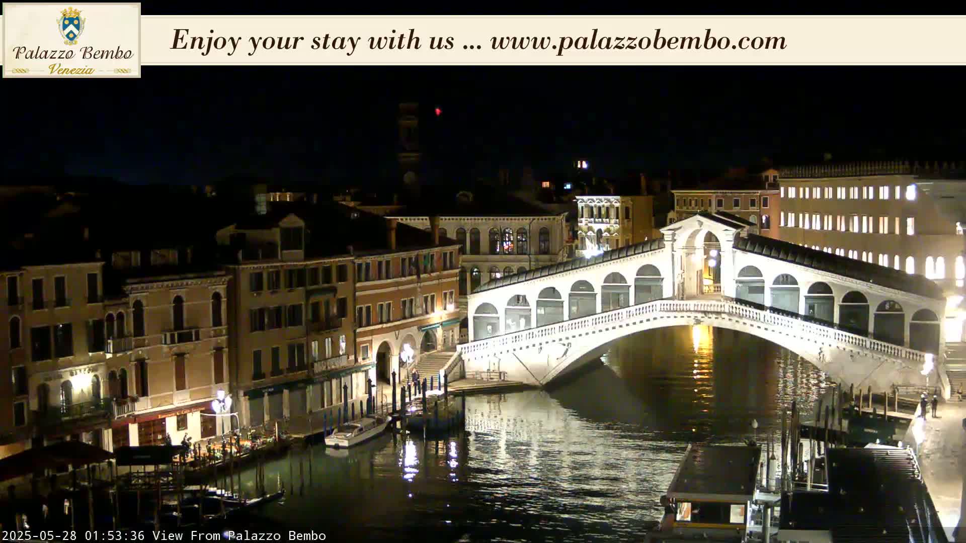 A brightly lit bridge spans a dark canal at night, with buildings lining the waterway.