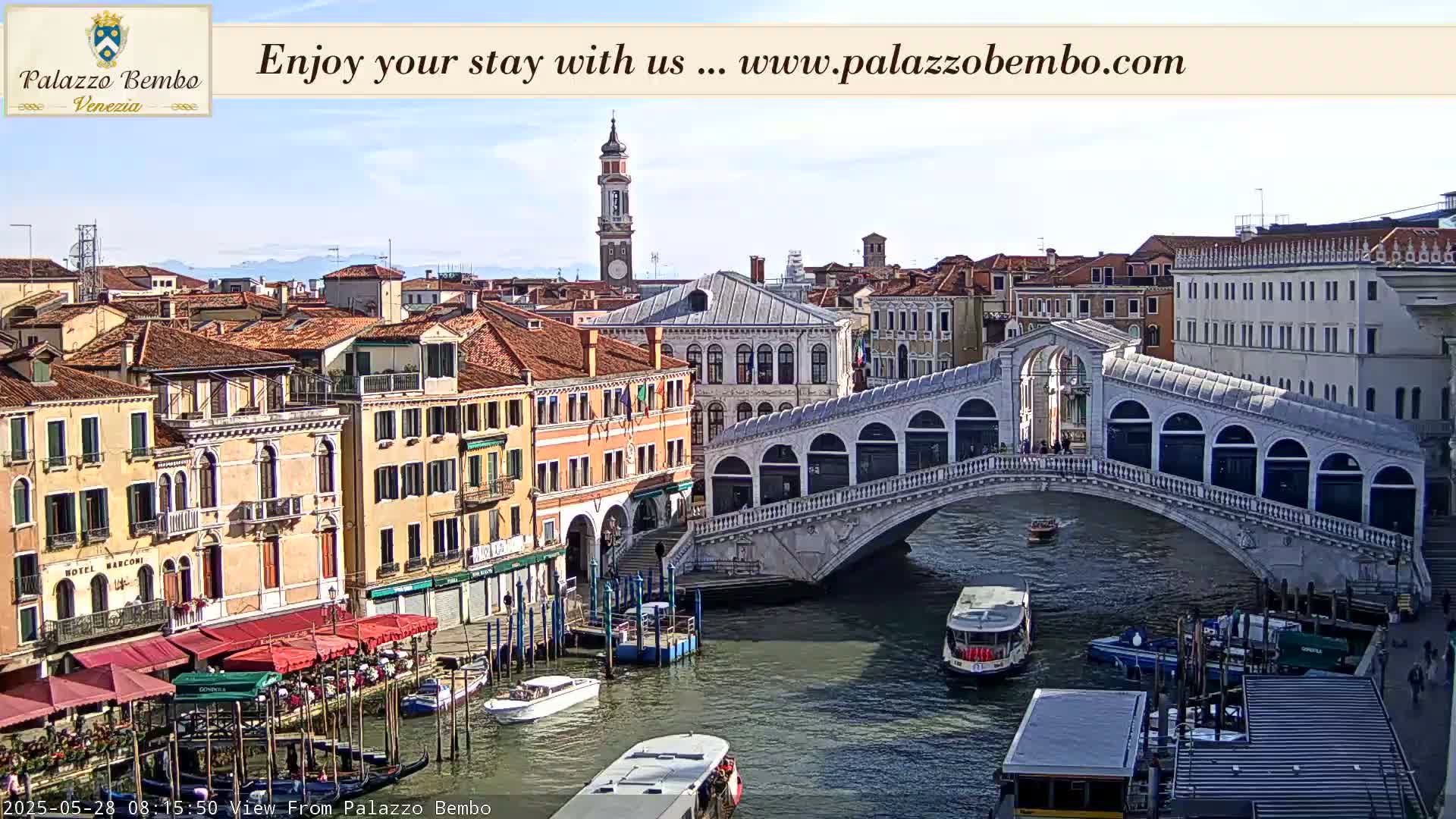 A sunny view of a canal in Venice, Italy, shows a bridge with people walking across it, boats on the water, and colorful buildings lining the canal.