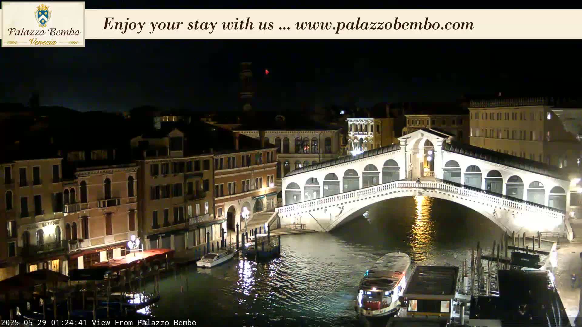 A nighttime view of a lit bridge over a canal in Venice, with boats moored along the sides of the buildings.