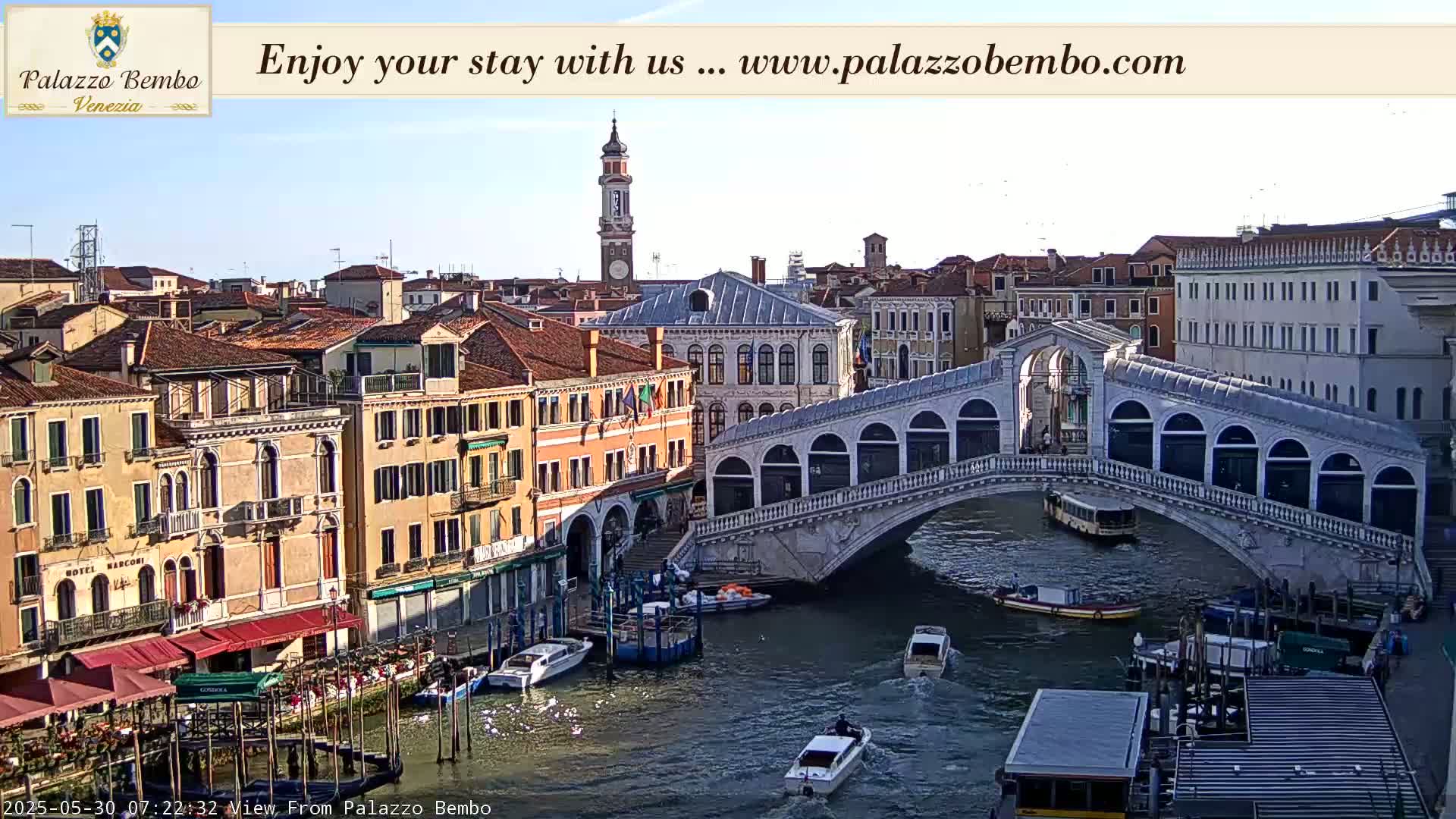 A sunny view of a canal in Venice, showing a bridge with pedestrians and boats on the water, surrounded by historic buildings.