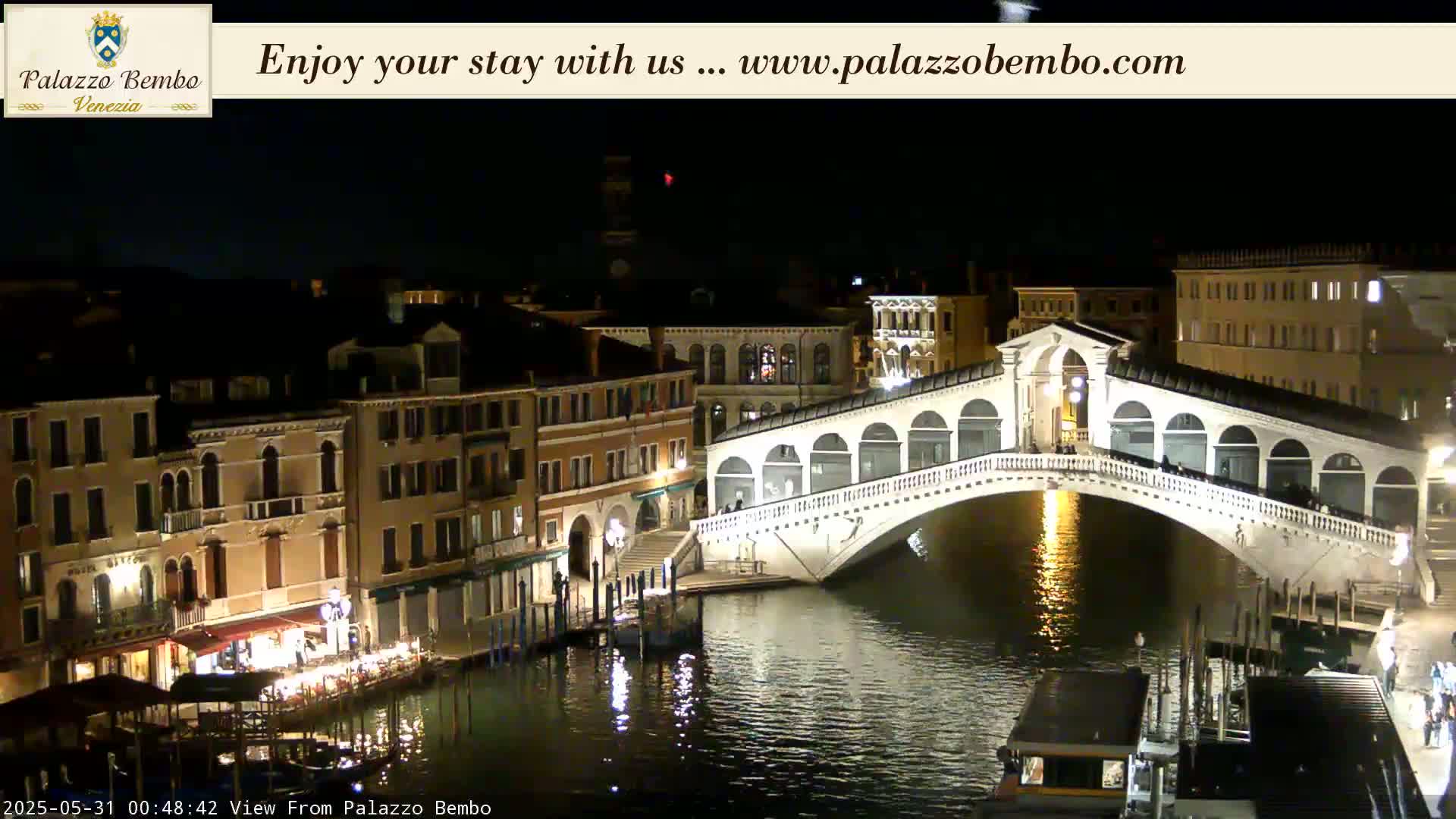 A nighttime view of a lit bridge spanning a canal in Venice, with buildings lining the waterway on either side.