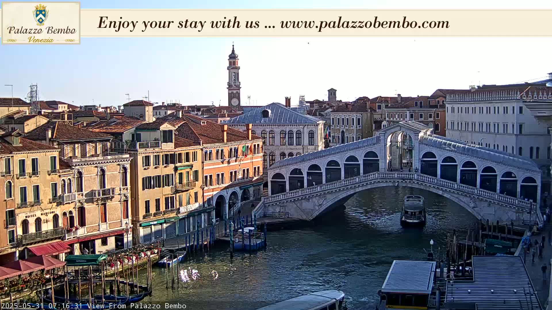 A Venetian canal scene under a clear sky shows a bridge with arched walkways, boats passing underneath, and colorful buildings lining the waterway.