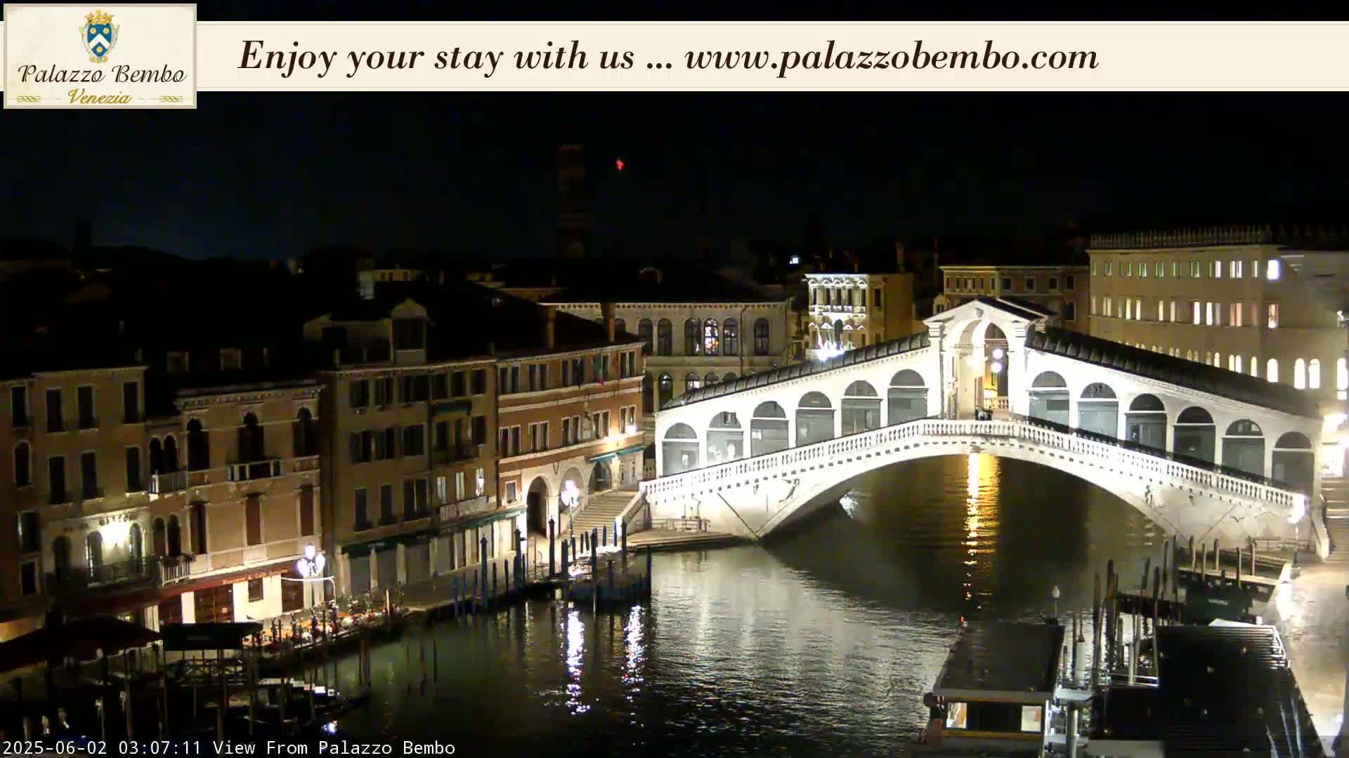 A nighttime view of a canal in Venice, showing the Rialto Bridge illuminated, with buildings lining the waterway on either side.