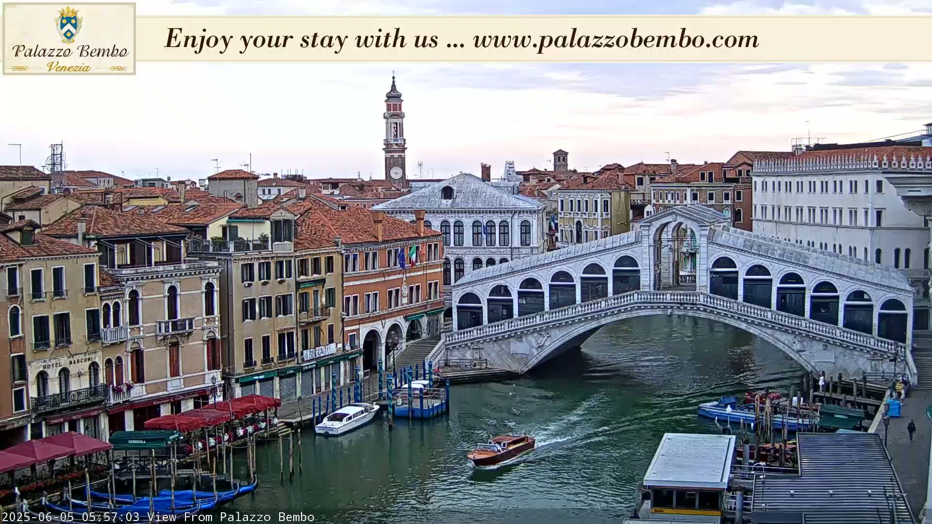 A Venetian canal scene under a partly cloudy sky shows a bridge arching over the water, with boats and buildings lining the canal.