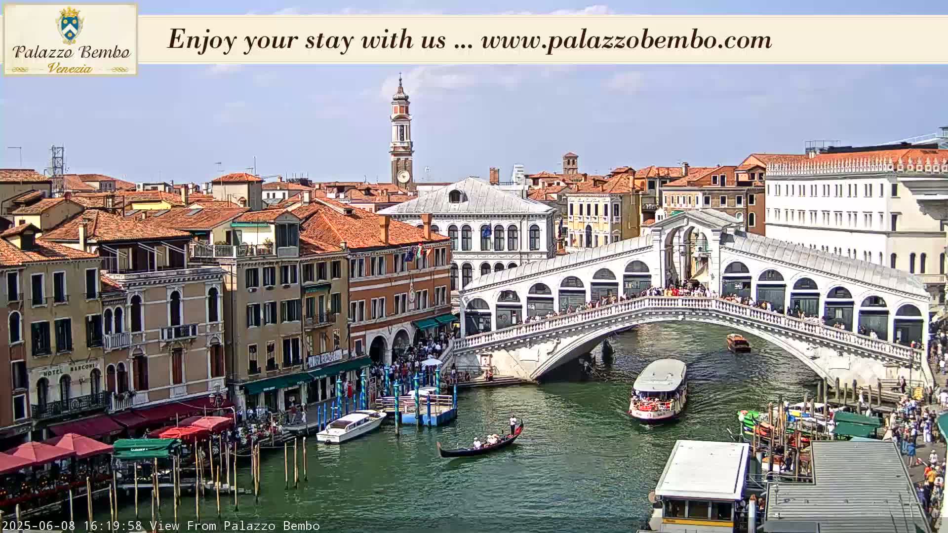 A sunny view of a canal in Venice, Italy, shows a bridge crowded with people and boats on the water.
