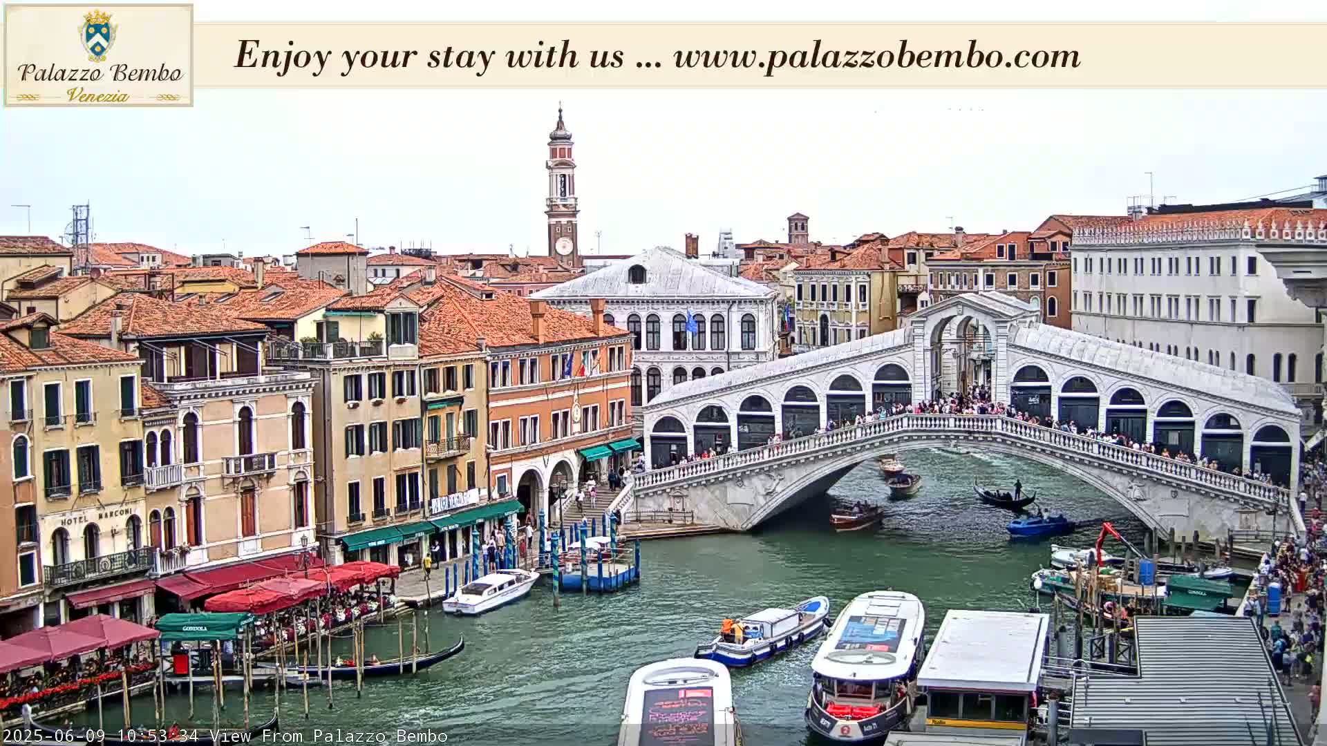 A canal in Venice, under a partly cloudy sky, is shown with a bridge crowded with people, boats, and buildings lining the waterway.