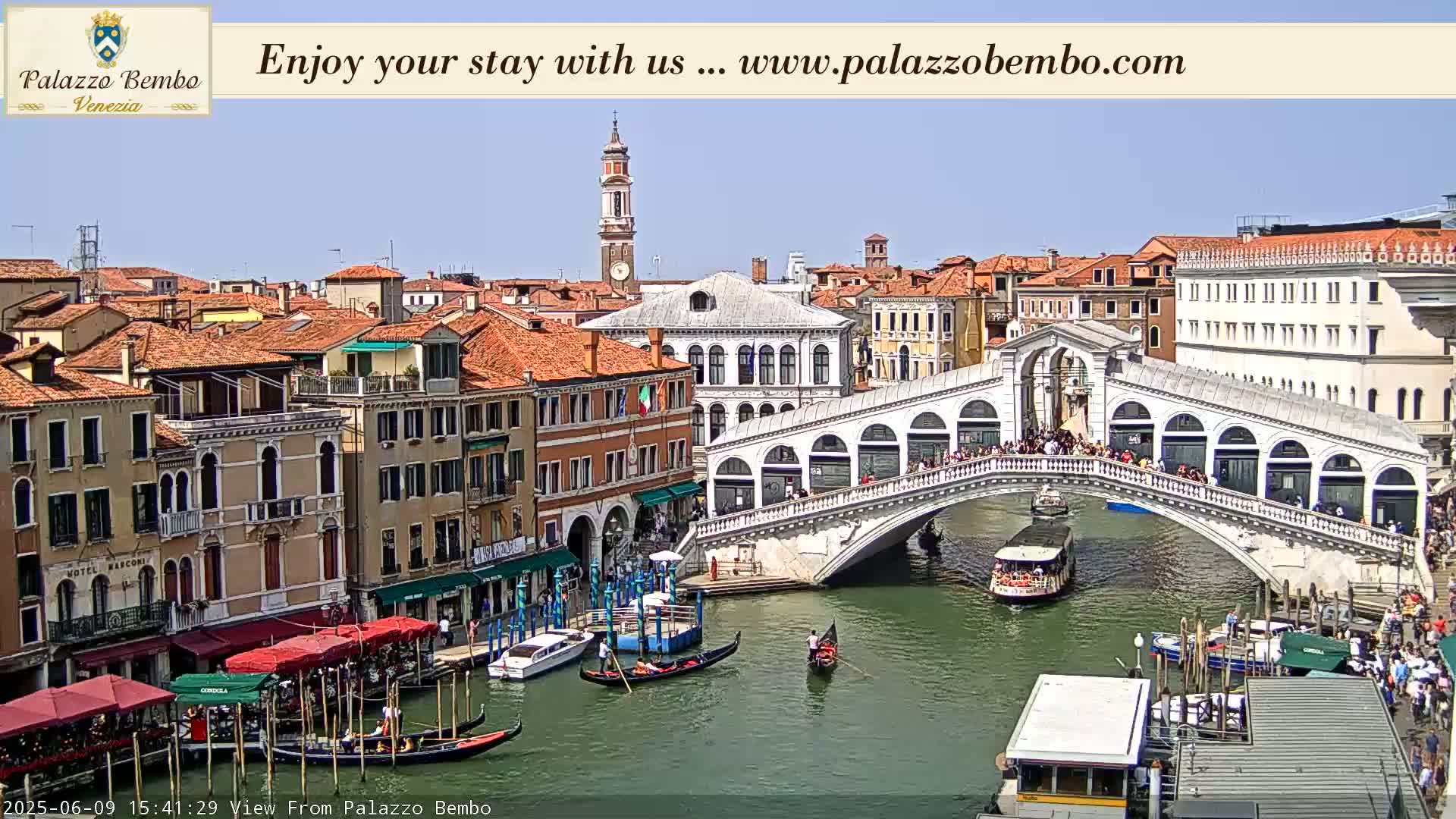 A sunny day in Venice shows the Rialto Bridge with many people crossing it and gondolas and boats on the canal below.