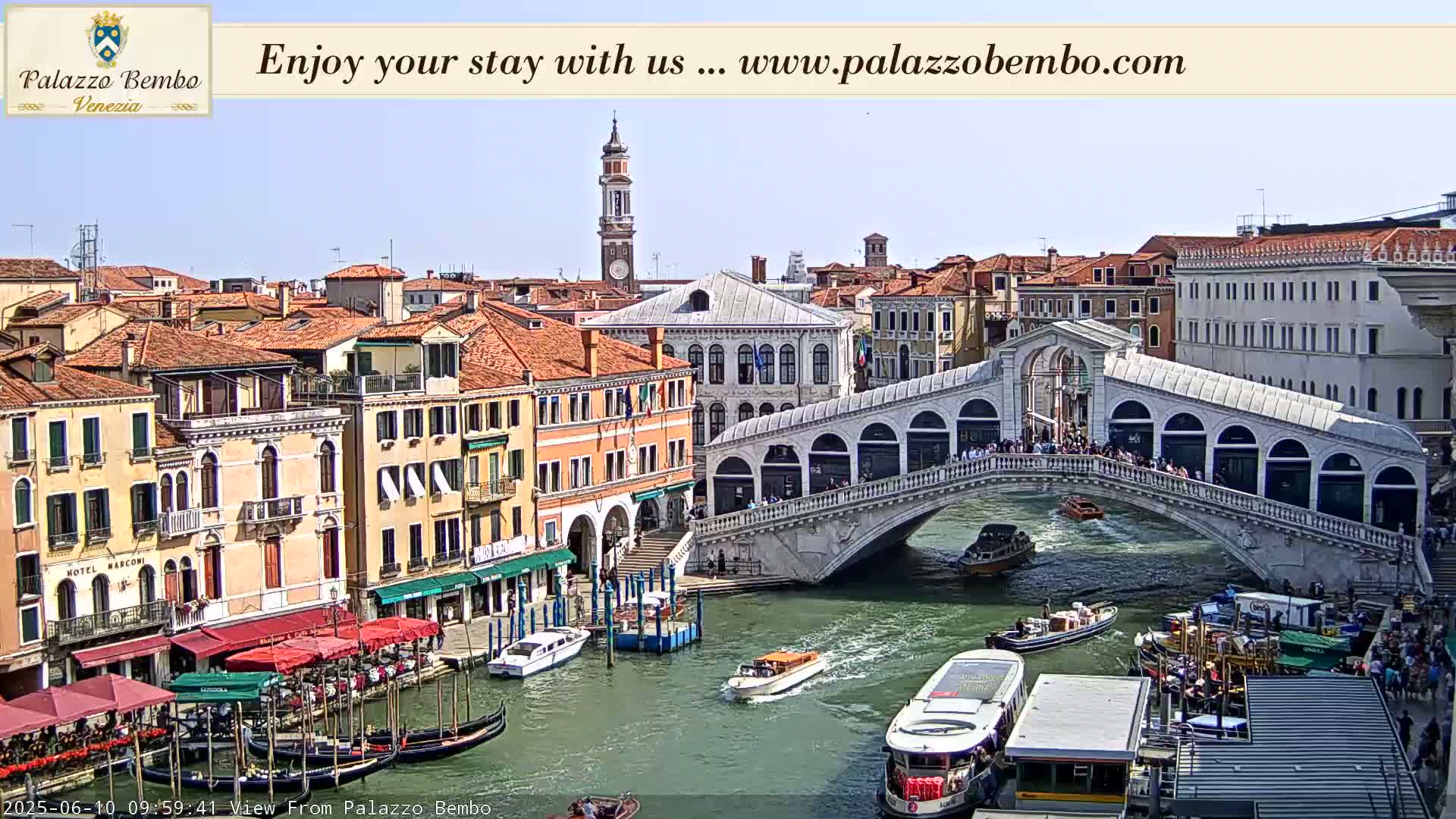 A sunny day in Venice shows a canal with numerous boats, gondolas, and the Rialto Bridge with people walking across it, and buildings lining the canal.