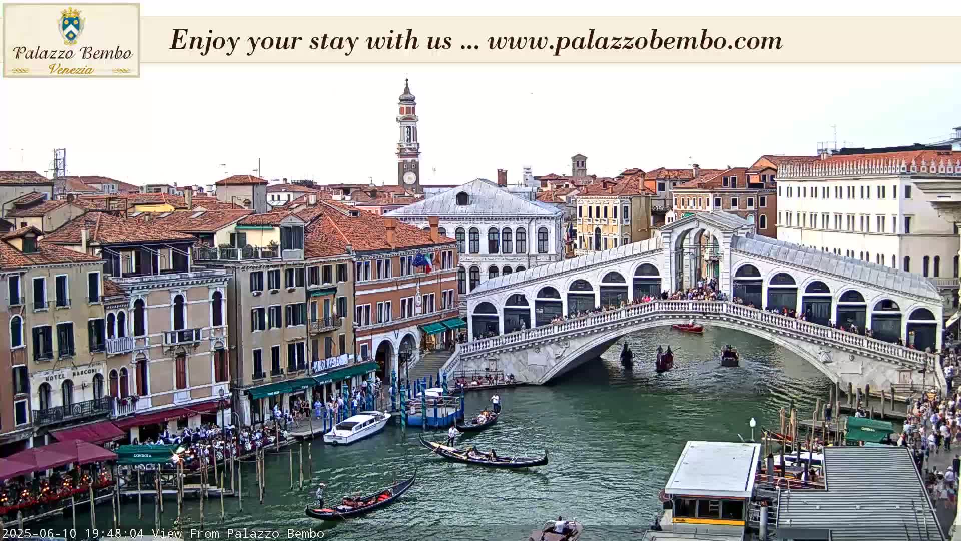 A sunny view of the Rialto Bridge in Venice, with gondolas and people on the bridge and along the canals below.