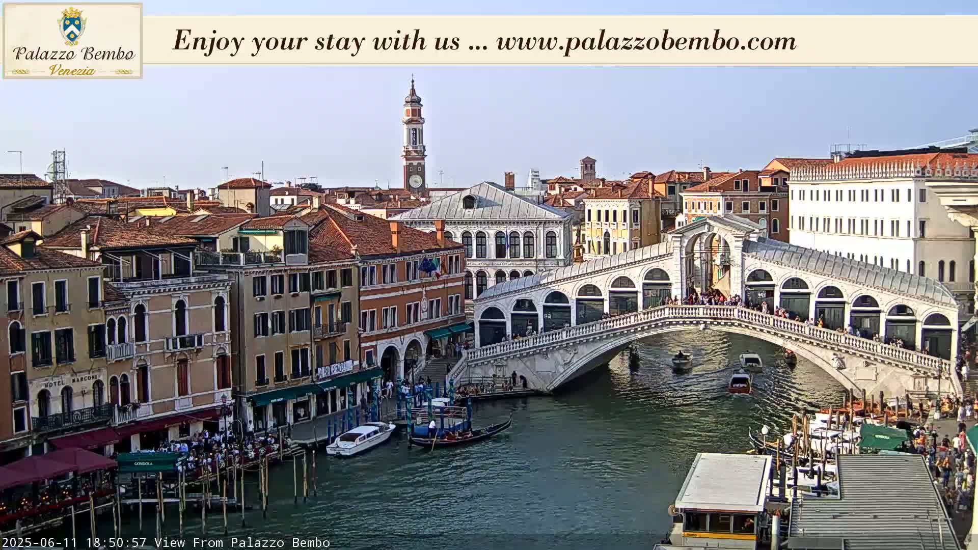 A sunny view of a Venetian canal with gondolas, boats, and the Rialto Bridge, showing many buildings and people.