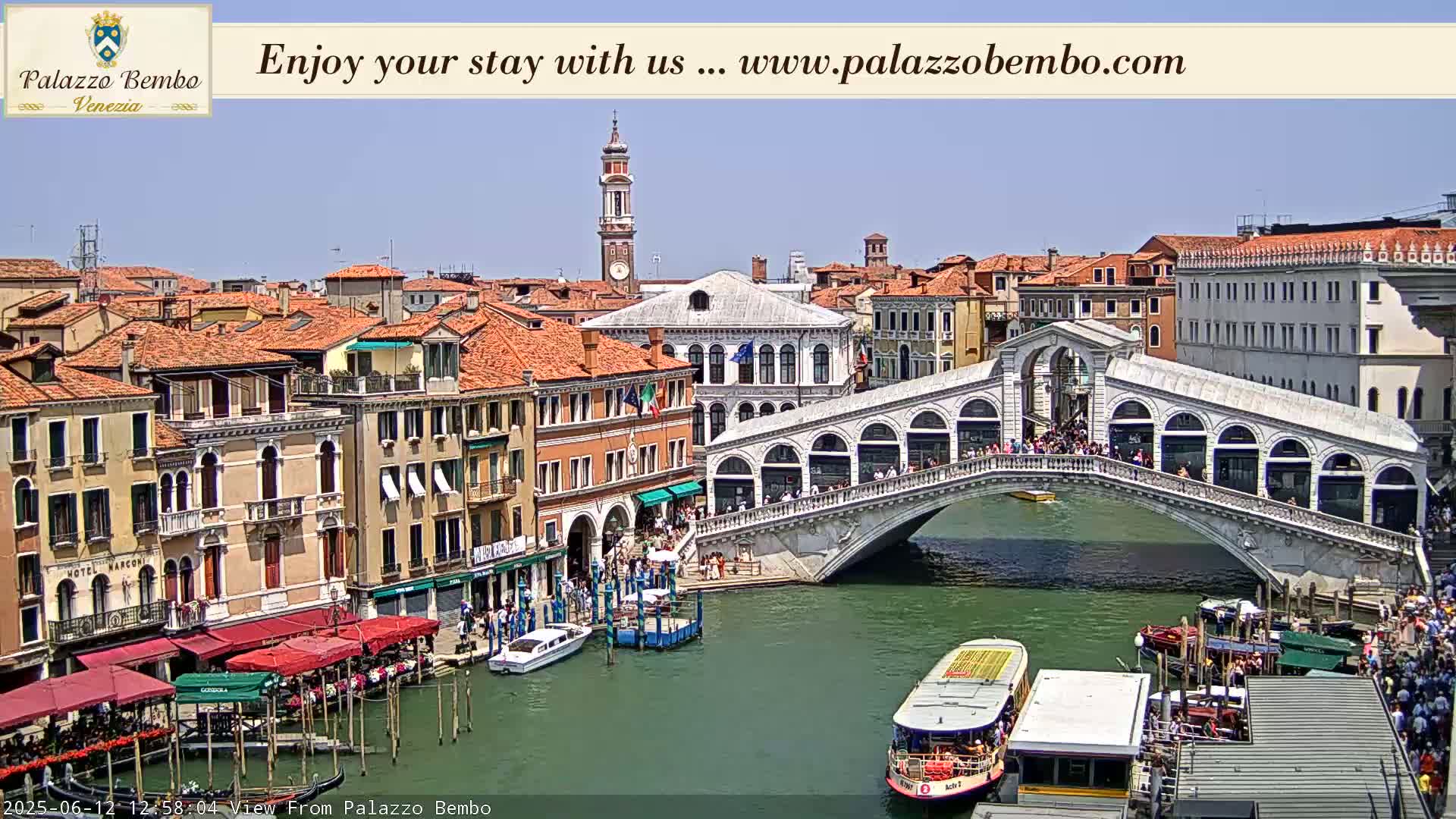 A sunny day in Venice shows a canal with boats, a bridge crowded with people, and numerous buildings with red-tiled roofs.