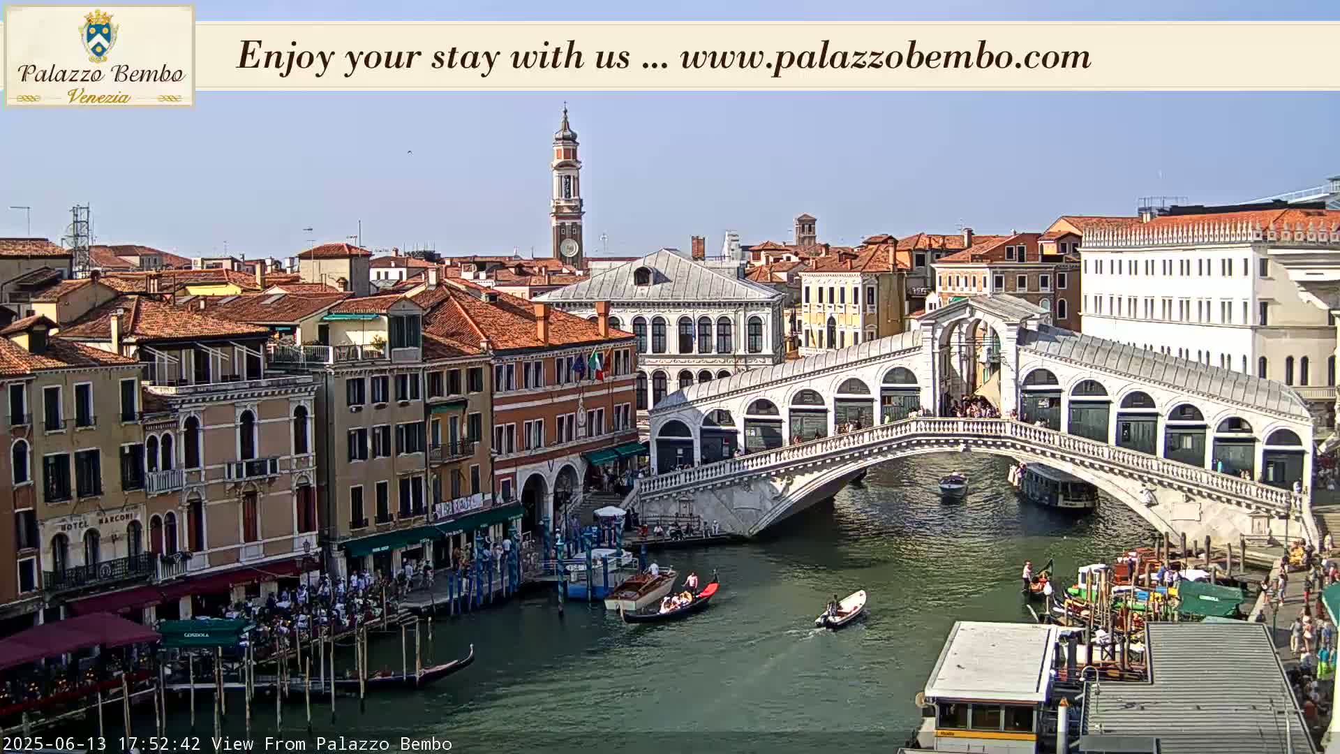 A sunny view of a canal in Venice, featuring the Rialto Bridge, gondolas, and numerous buildings along the waterway.