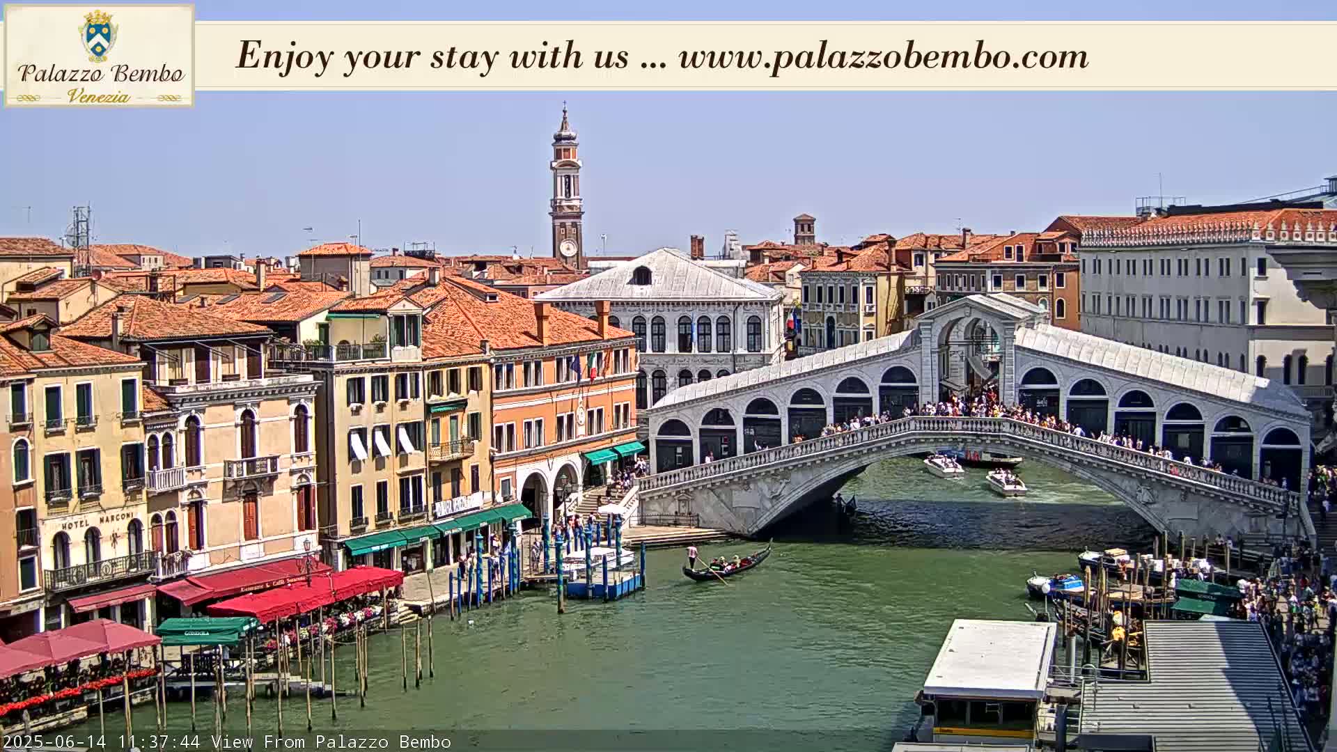 A sunny view of a canal in Venice, showing a bridge crowded with people and numerous boats, with buildings lining the waterway.