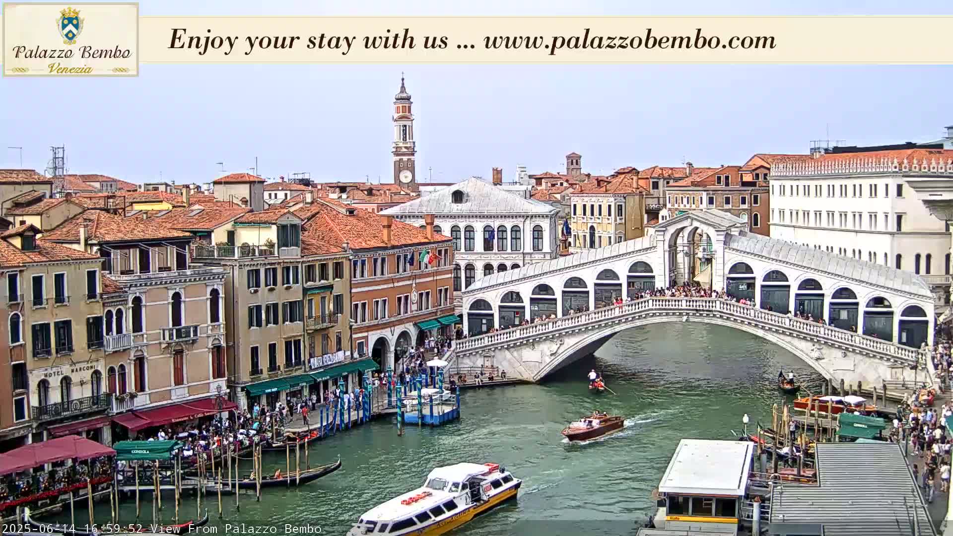 A sunny view of a canal in Venice, showing the Rialto Bridge crowded with people, gondolas, and water taxis.