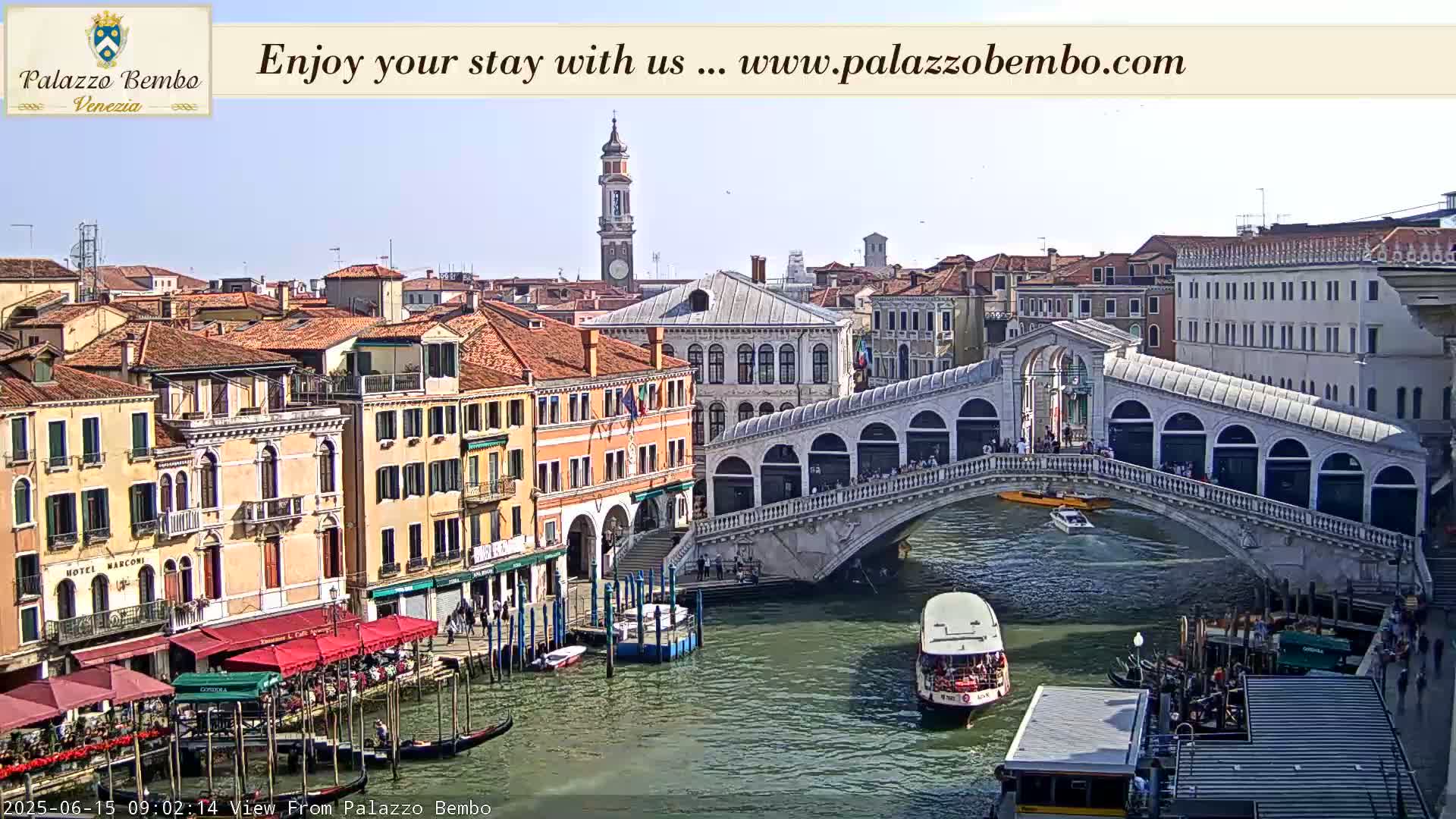 A sunny view of a canal in Venice, with boats passing under a bridge and buildings lining the waterway.