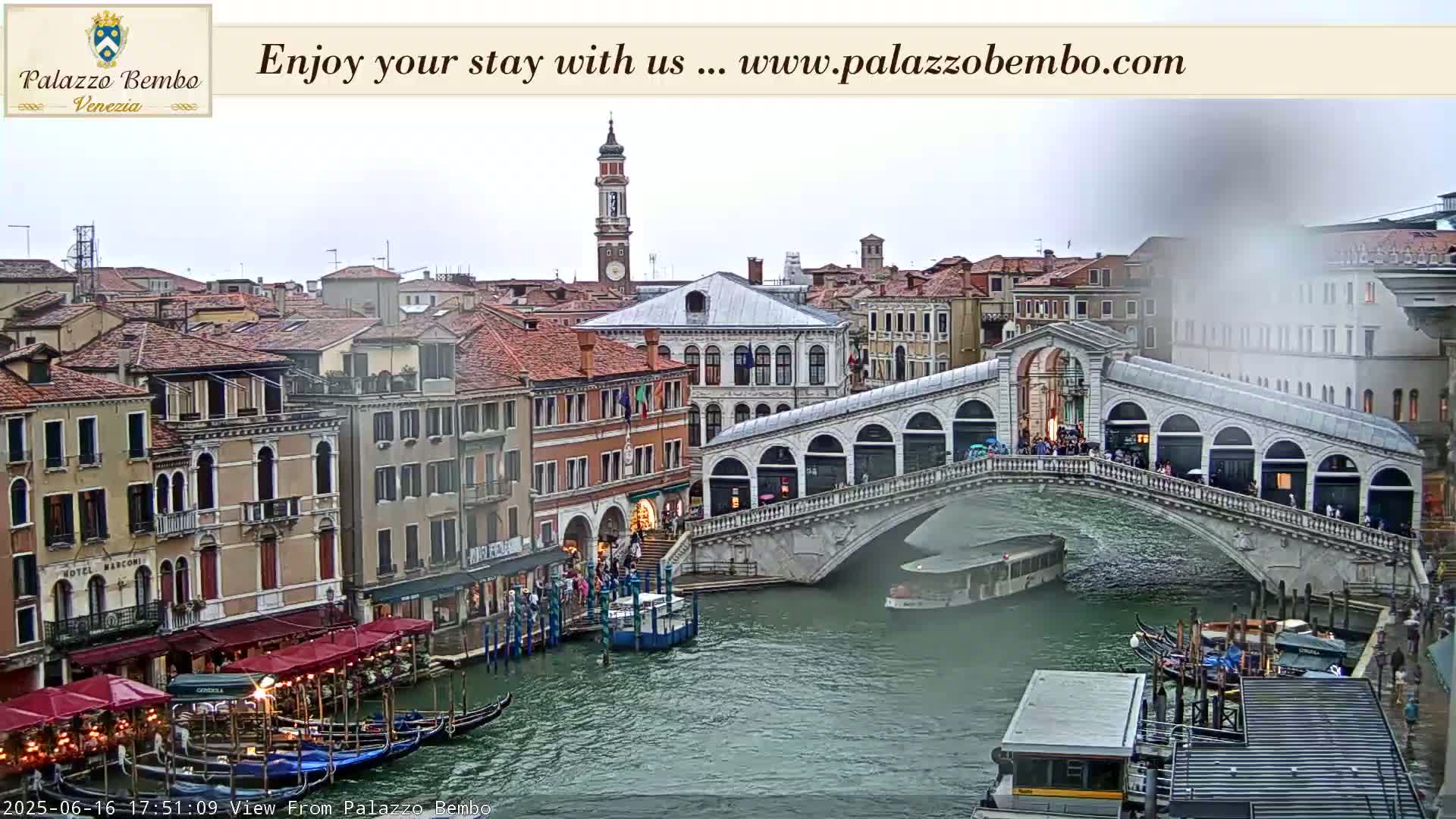 A view of a canal in Venice on an overcast day, showing gondolas, a bridge with people on it, and several buildings along the waterfront.